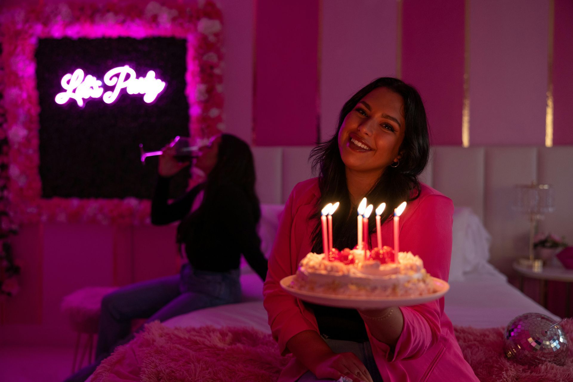 Woman holding a birthday cake with lit candles, smiling in a pink-themed room. Another person is in the background. 