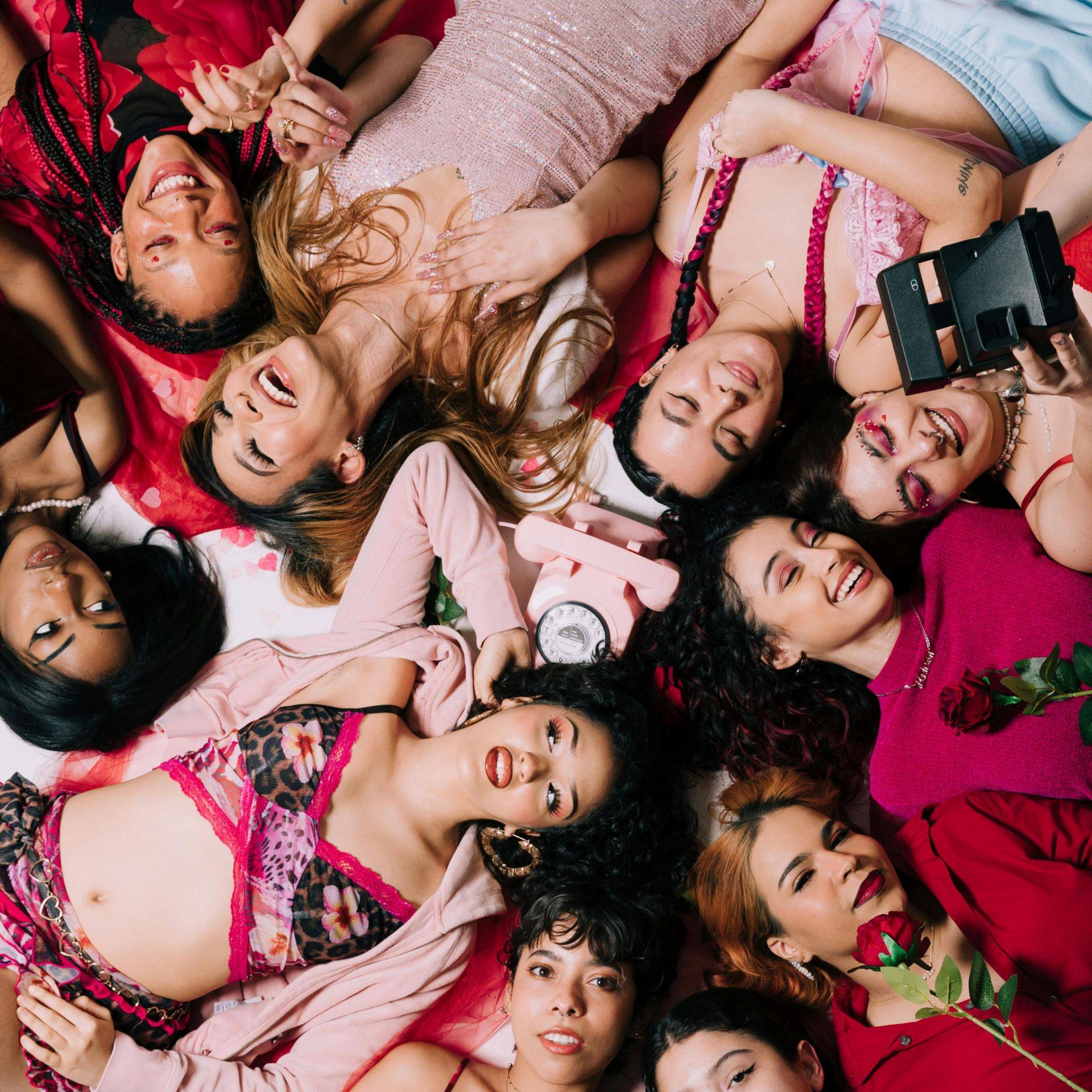 Eine Gruppe lächelnder Frauen liegt auf einer mit roten und rosa Stoffen bedeckten Fläche. Einige halten Gegenstände in den Händen, darunter eine Rose und ein Telefon.
