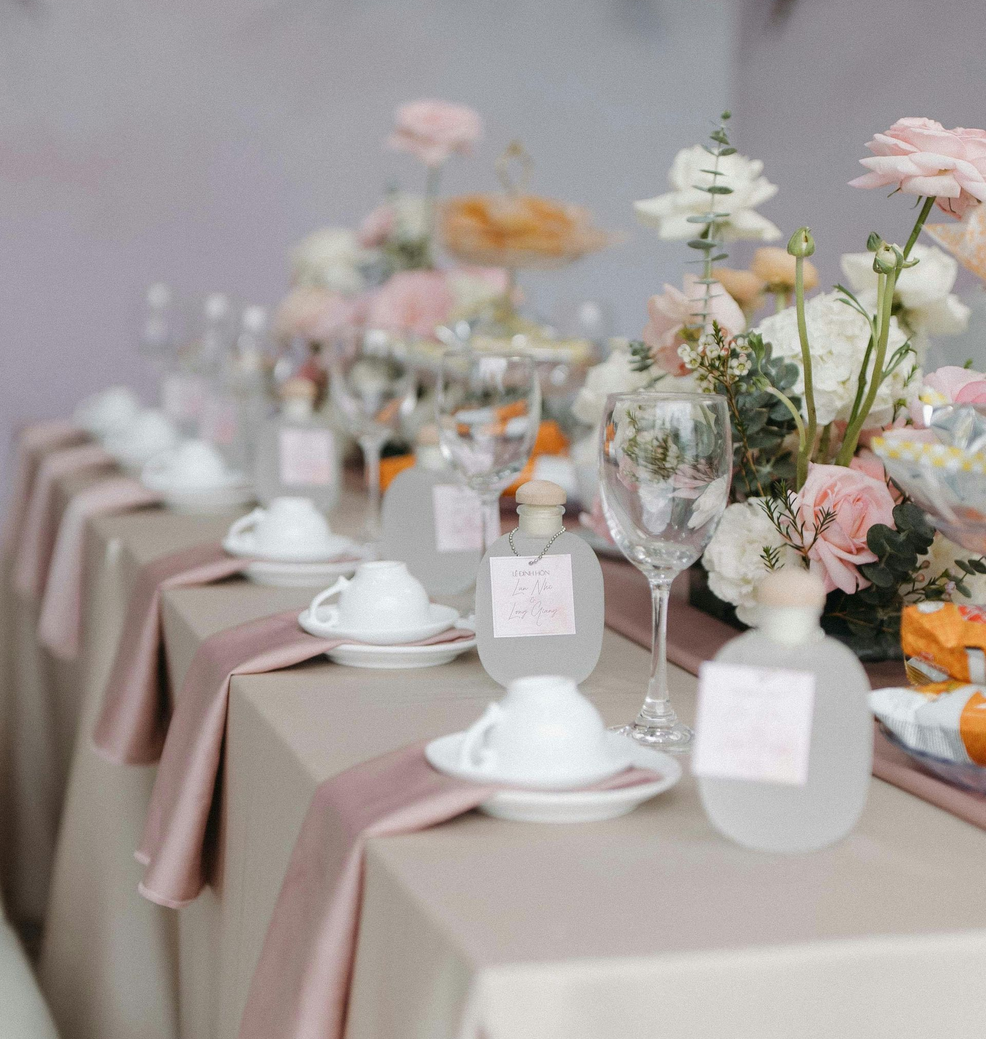 Gedeckter Tisch mit Teetassen, Weingläsern und Blumenarrangements. Beige- und Rosatöne.