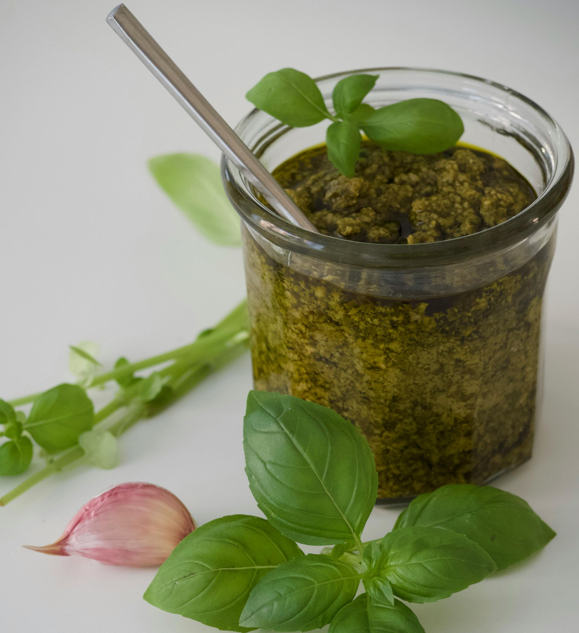 Ein Glas Pesto mit Basilikumblättern, Knoblauch und einem Löffel auf einer weißen Oberfläche.