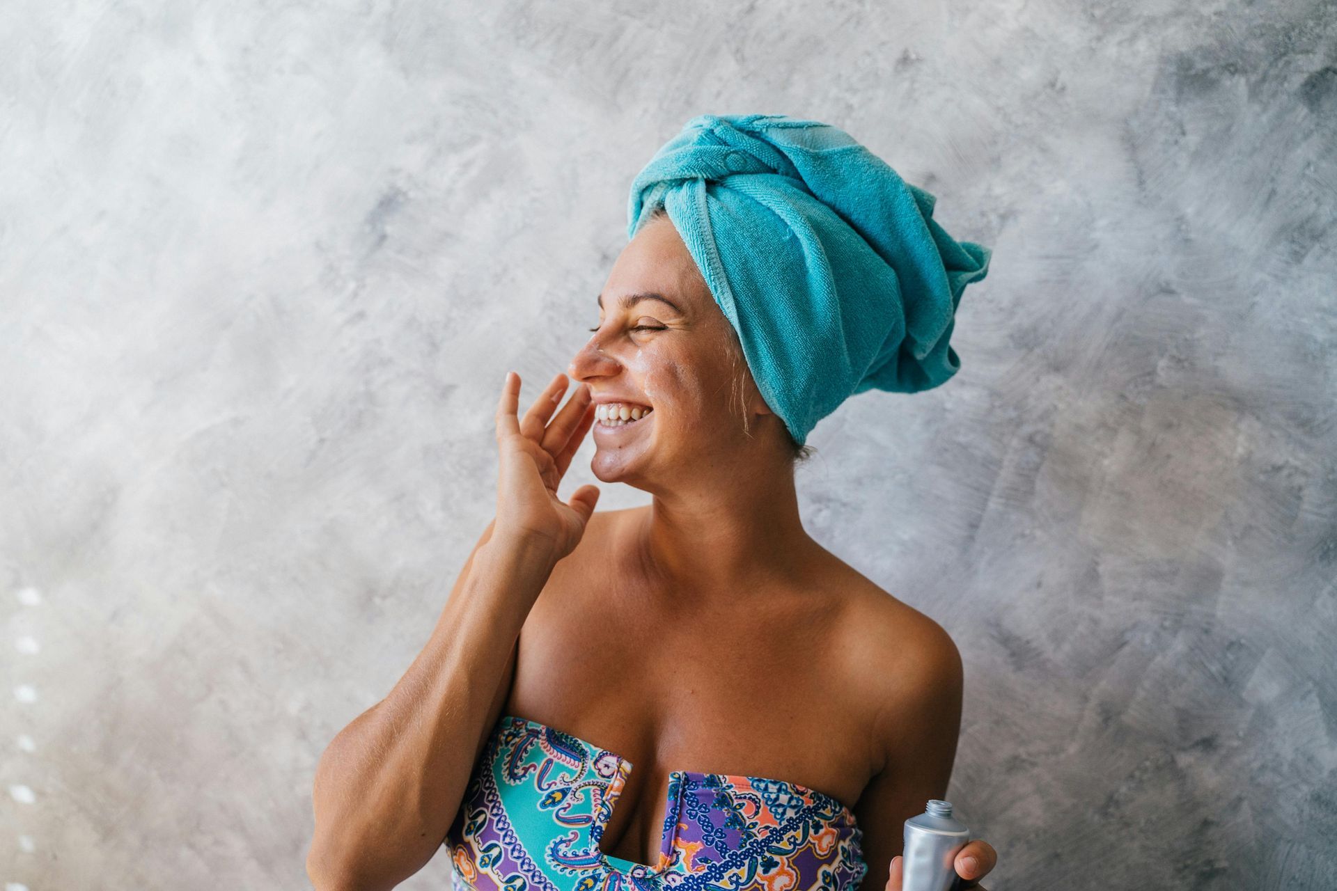 Smiling woman with a turquoise towel on her head, wearing a colorful strapless top, laughing against a gray wall