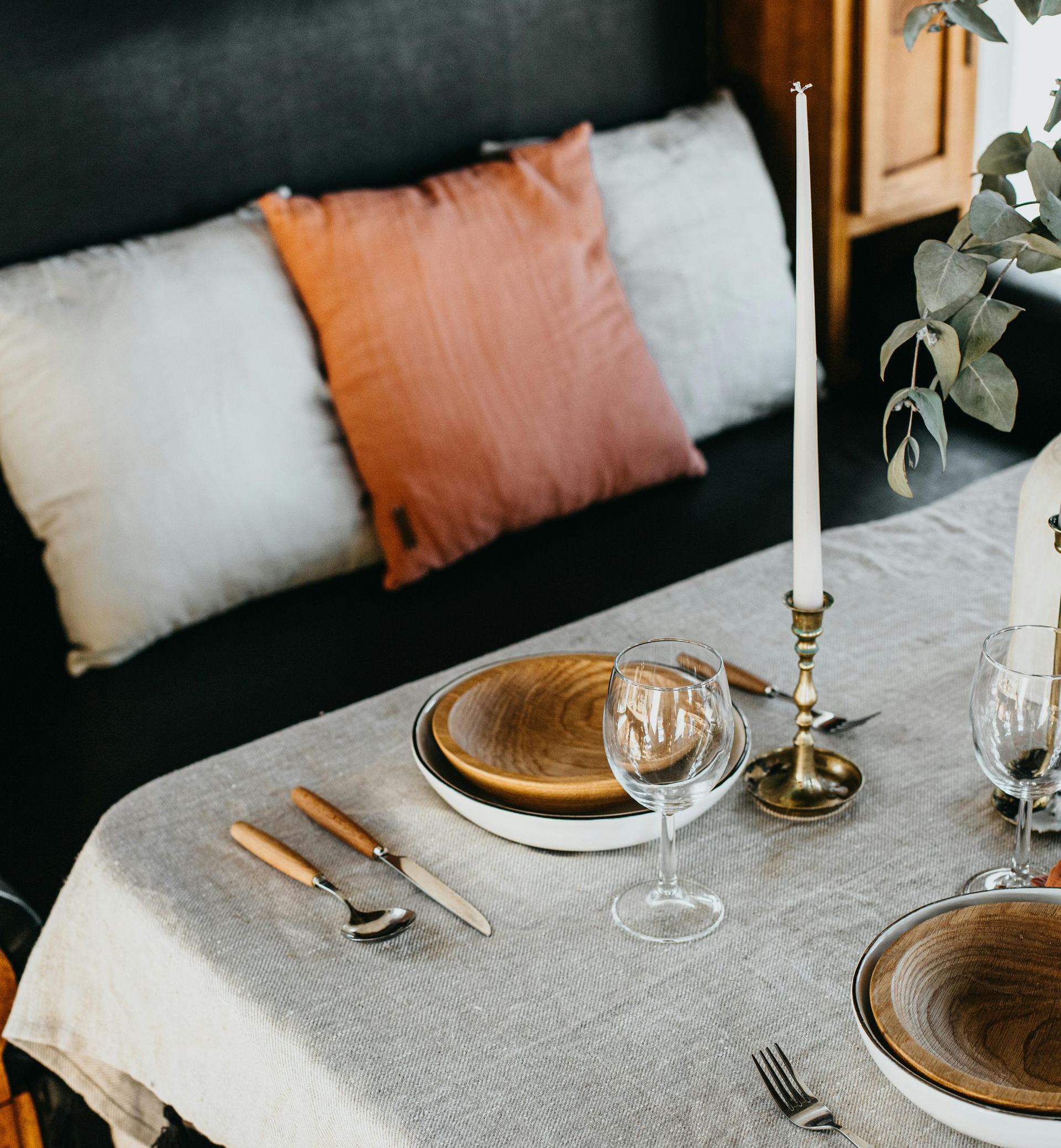 Gedeckter Tisch mit Tellern, Besteck und einer brennenden Kerze. Im Hintergrund Kissen auf einem dunklen Sofa.