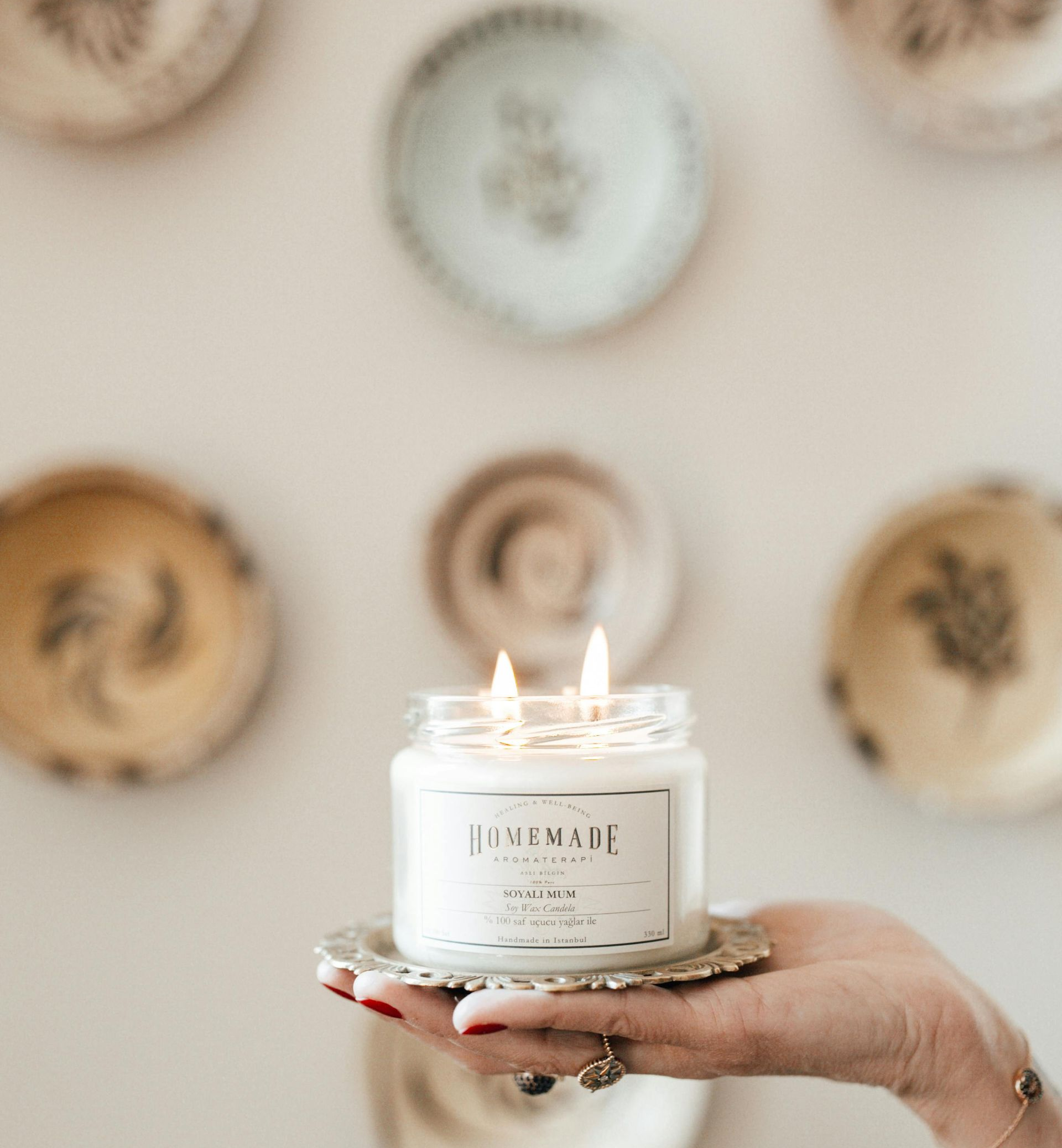 Eine Hand hält eine brennende Kerze auf einem dekorativen Teller; im Hintergrund sind rustikale Teller an einer weißen Wand zu sehen.