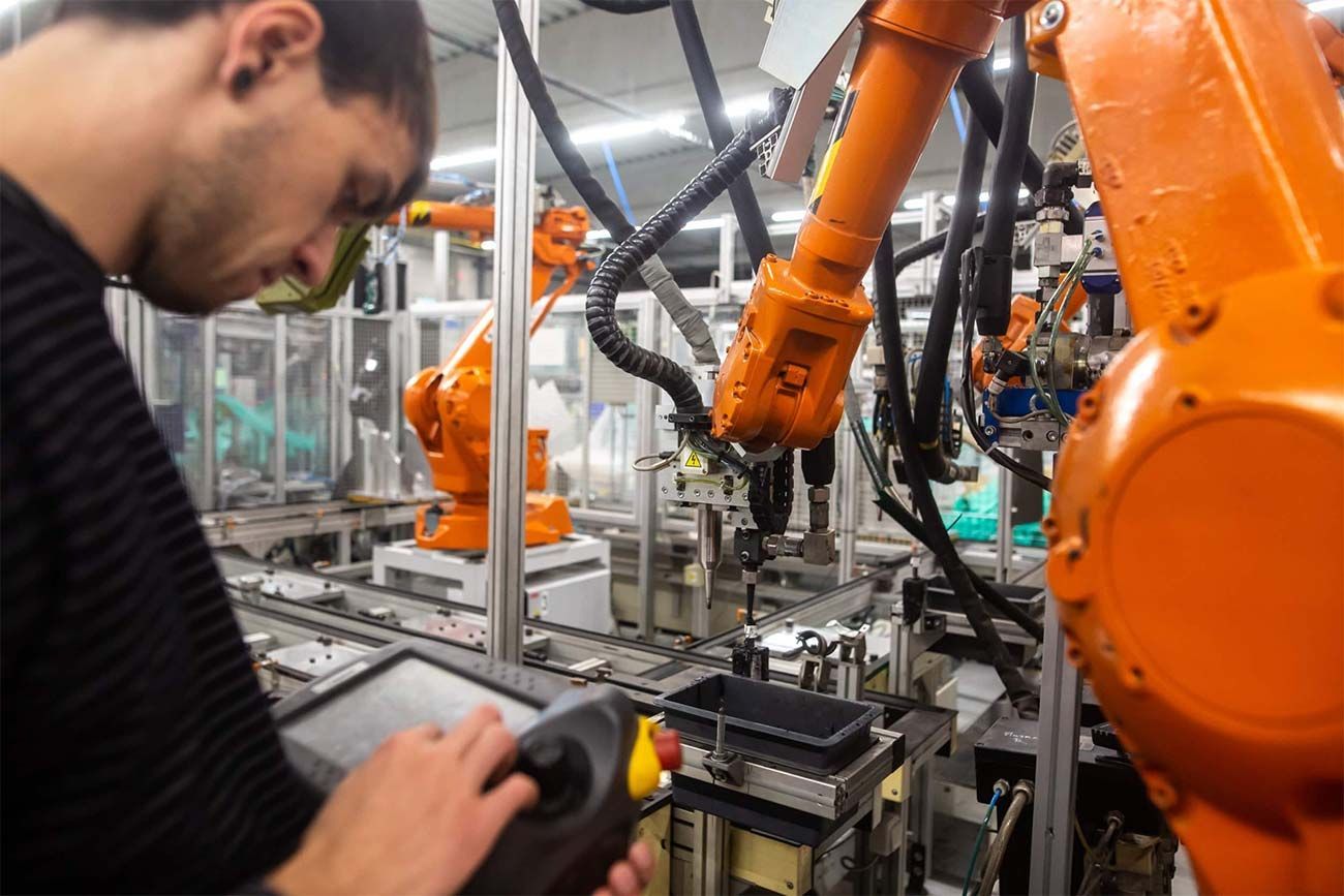 Un trabajador maneja un dispositivo de control junto a brazos robóticos industriales de color naranja en una fábrica.