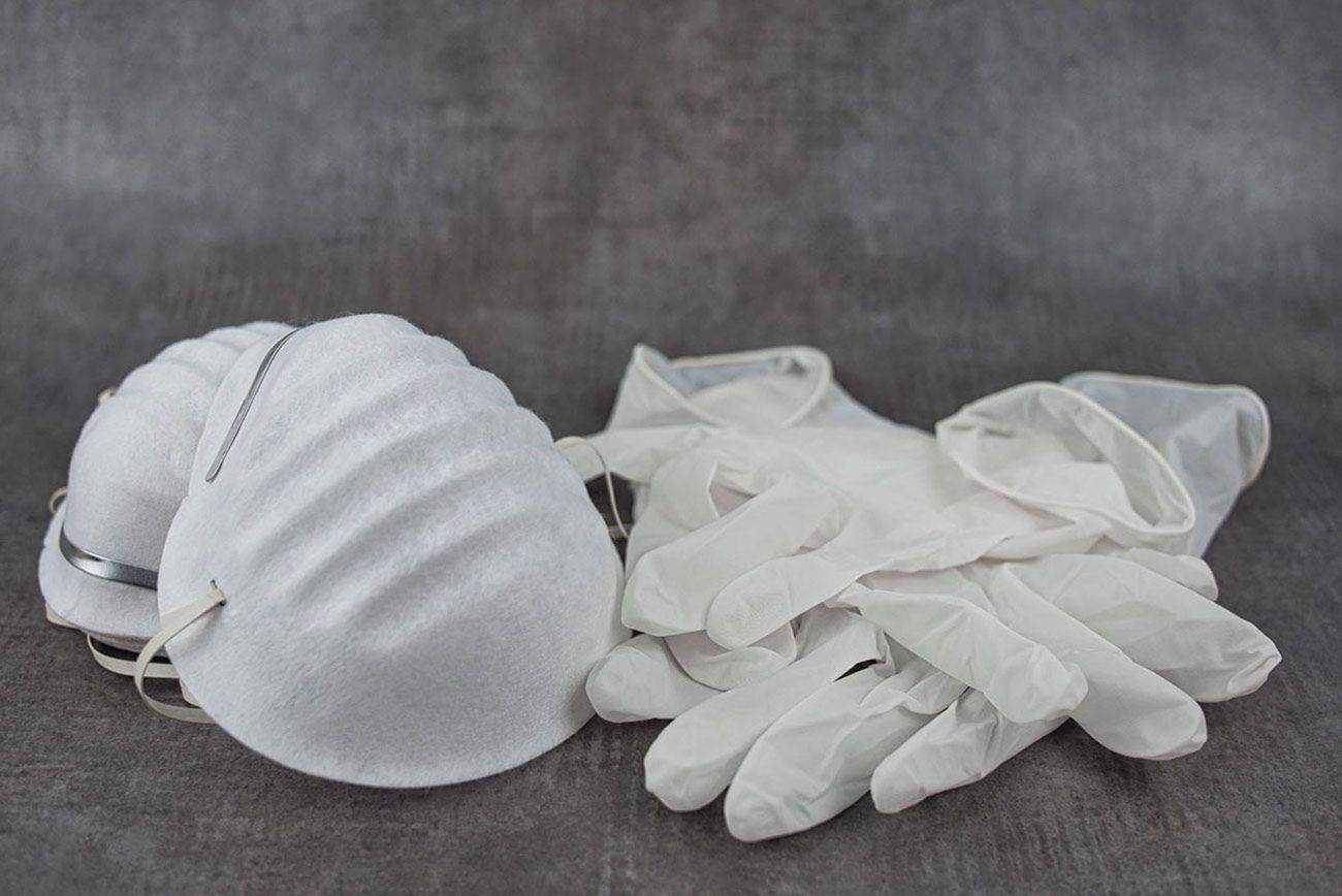 Dos mascarillas quirúrgicas blancas junto a un par de guantes blancos sobre una superficie gris.