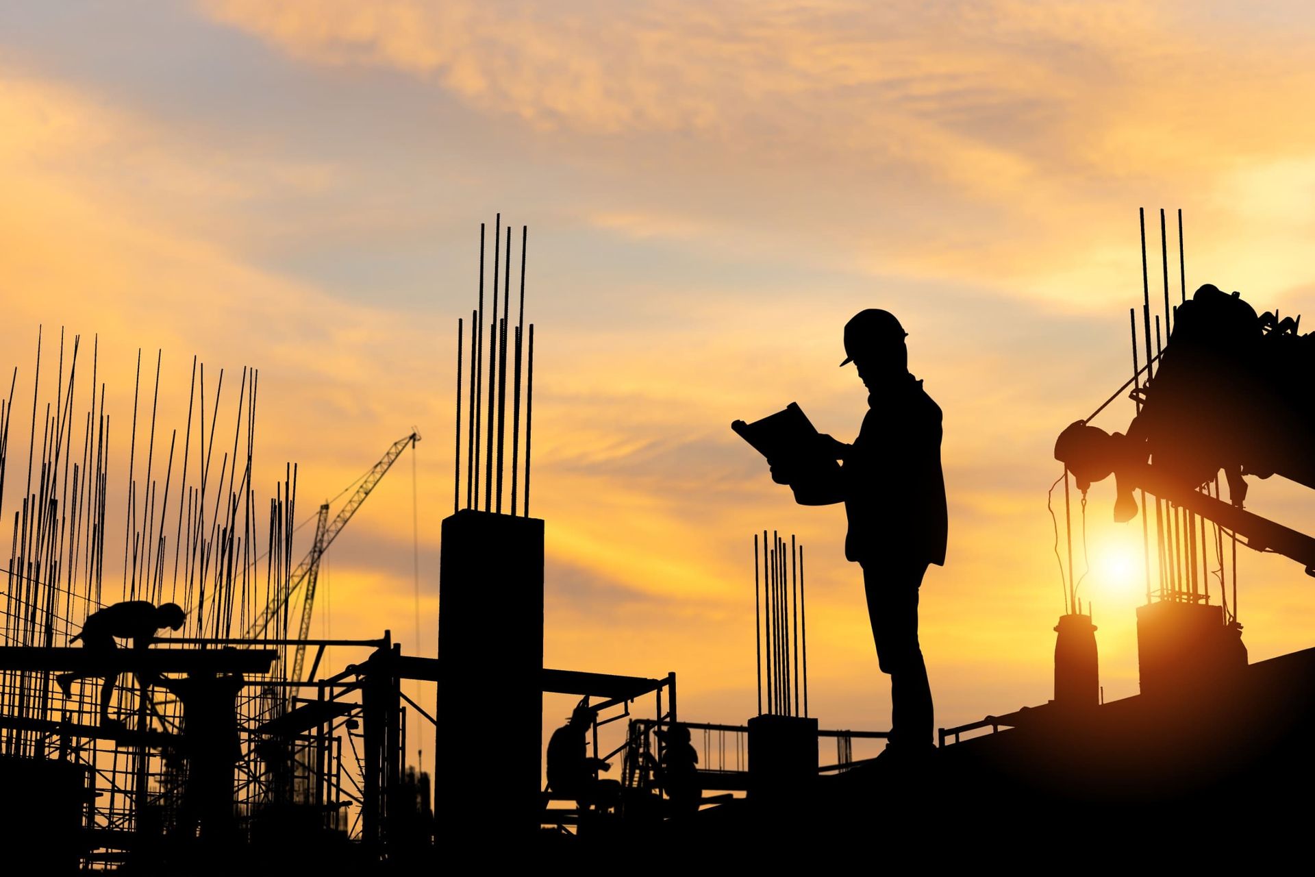 Trabajadores en silueta en una obra de construcción al atardecer, con barras de refuerzo y equipos contra el cielo anaranjado.