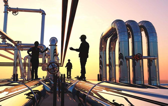 Trabajadores silueteados en una plataforma industrial con tuberías brillantes al atardecer.