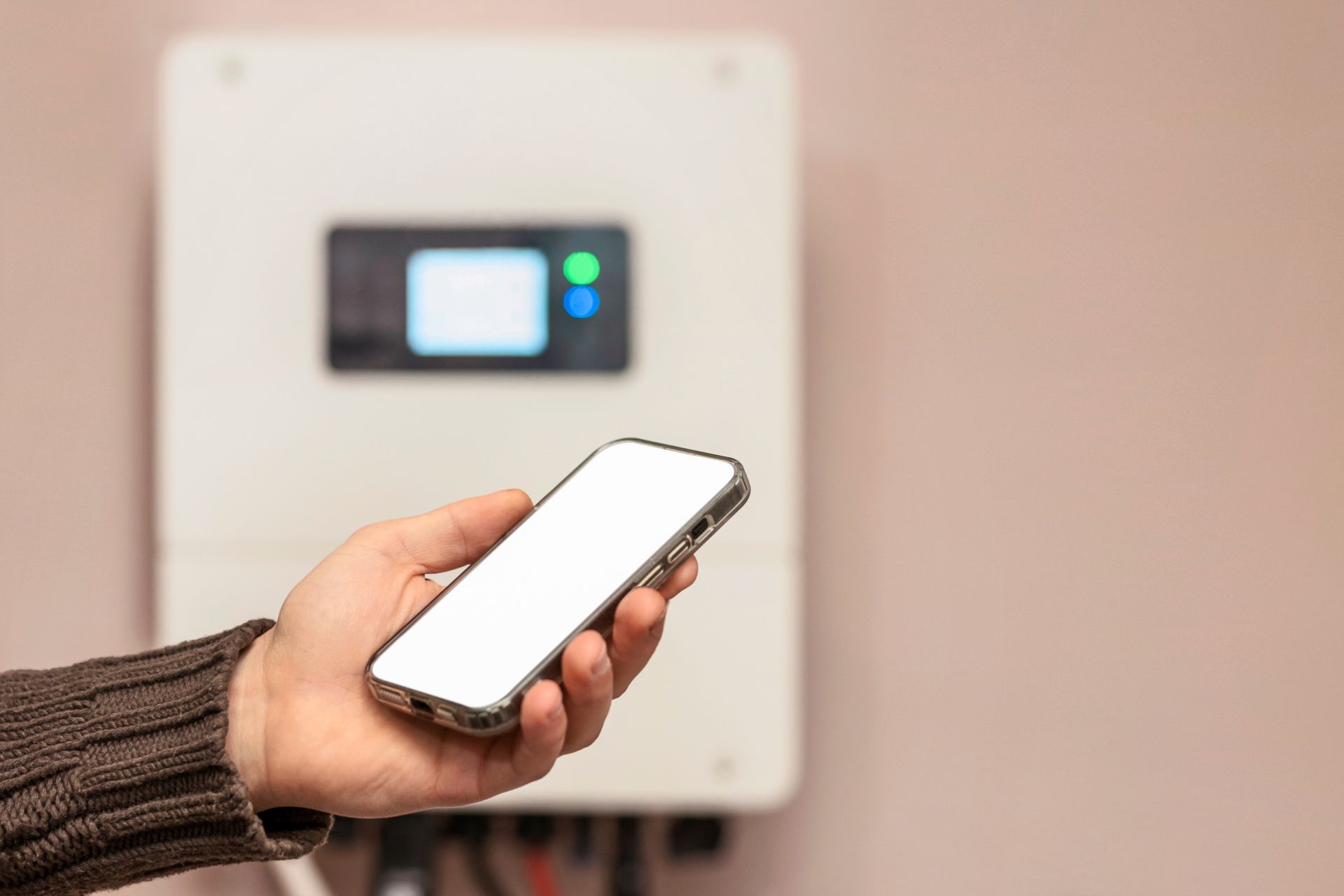 Une personne tient un smartphone devant un onduleur solaire blanc fixé au mur.