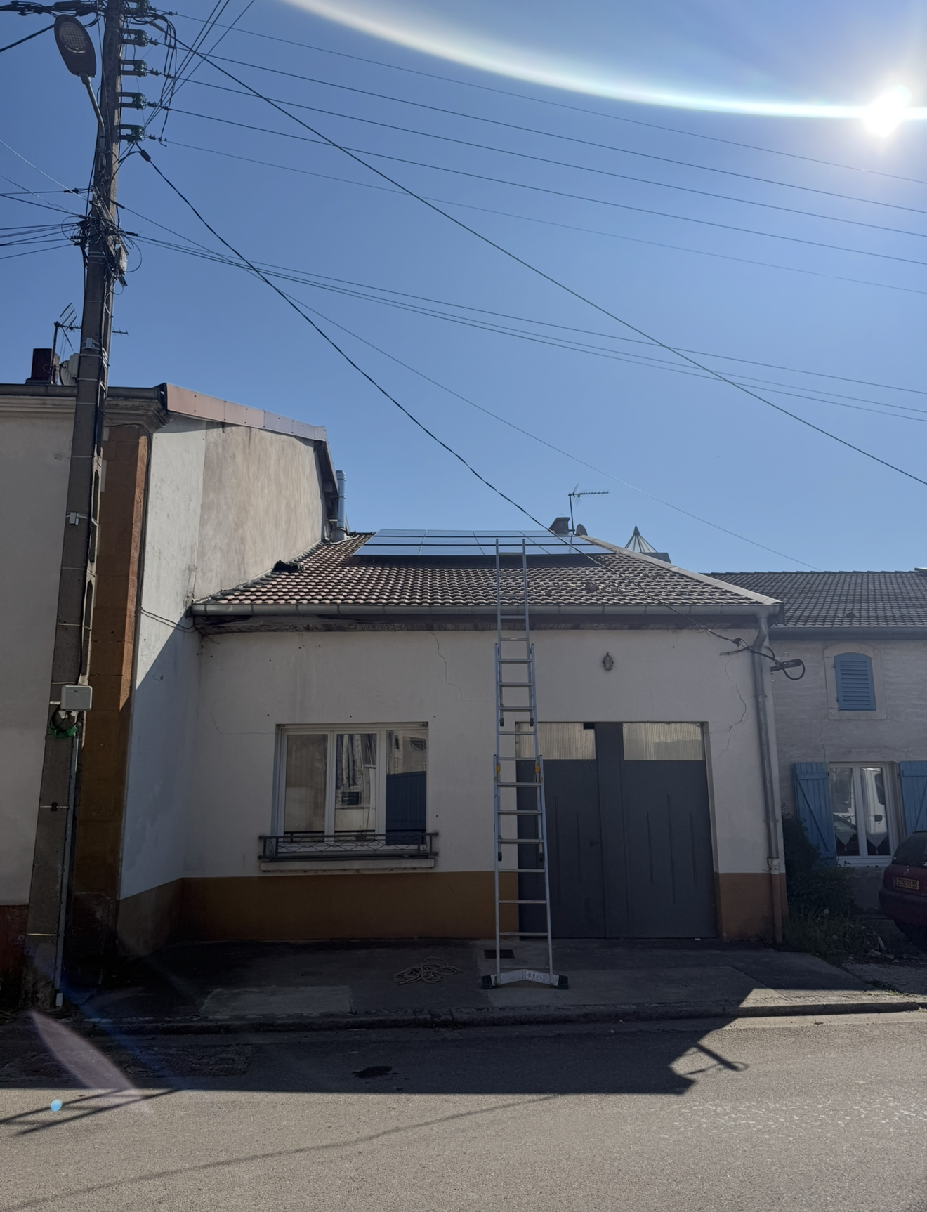 Une échelle est appuyée contre la façade blanche d'une petite maison dont le toit est recouvert de panneaux solaires.