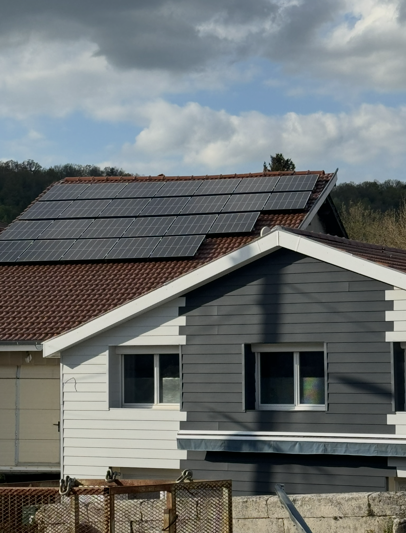 Une maison au toit recouvert de panneaux solaires, avec des murs extérieurs blancs et gris foncé sous un ciel nuageux.