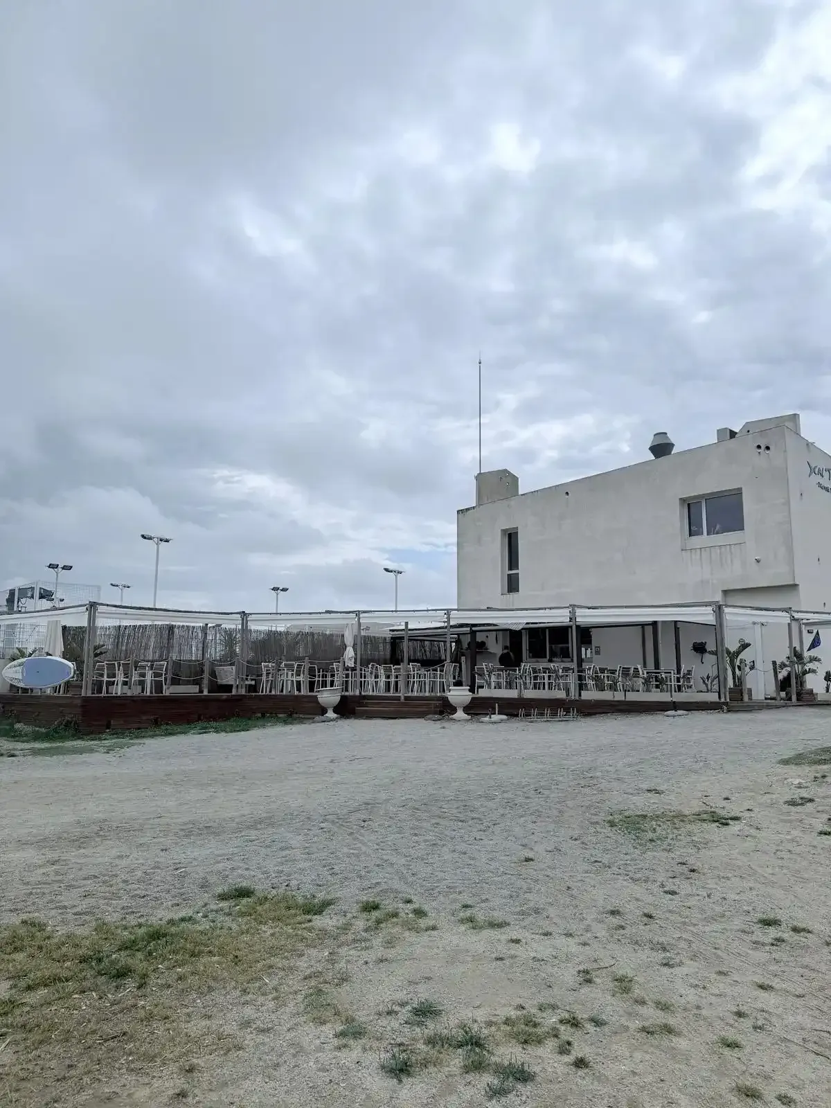 Terreno de grava junto a un edificio blanco de dos plantas bajo un cielo nublado, con un largo pasillo cubierto.