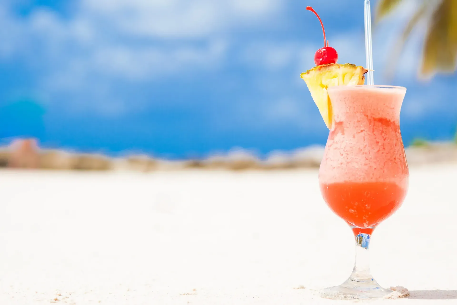 Cóctel tropical rosa con piña y cereza en una playa, con el cielo azul de fondo.