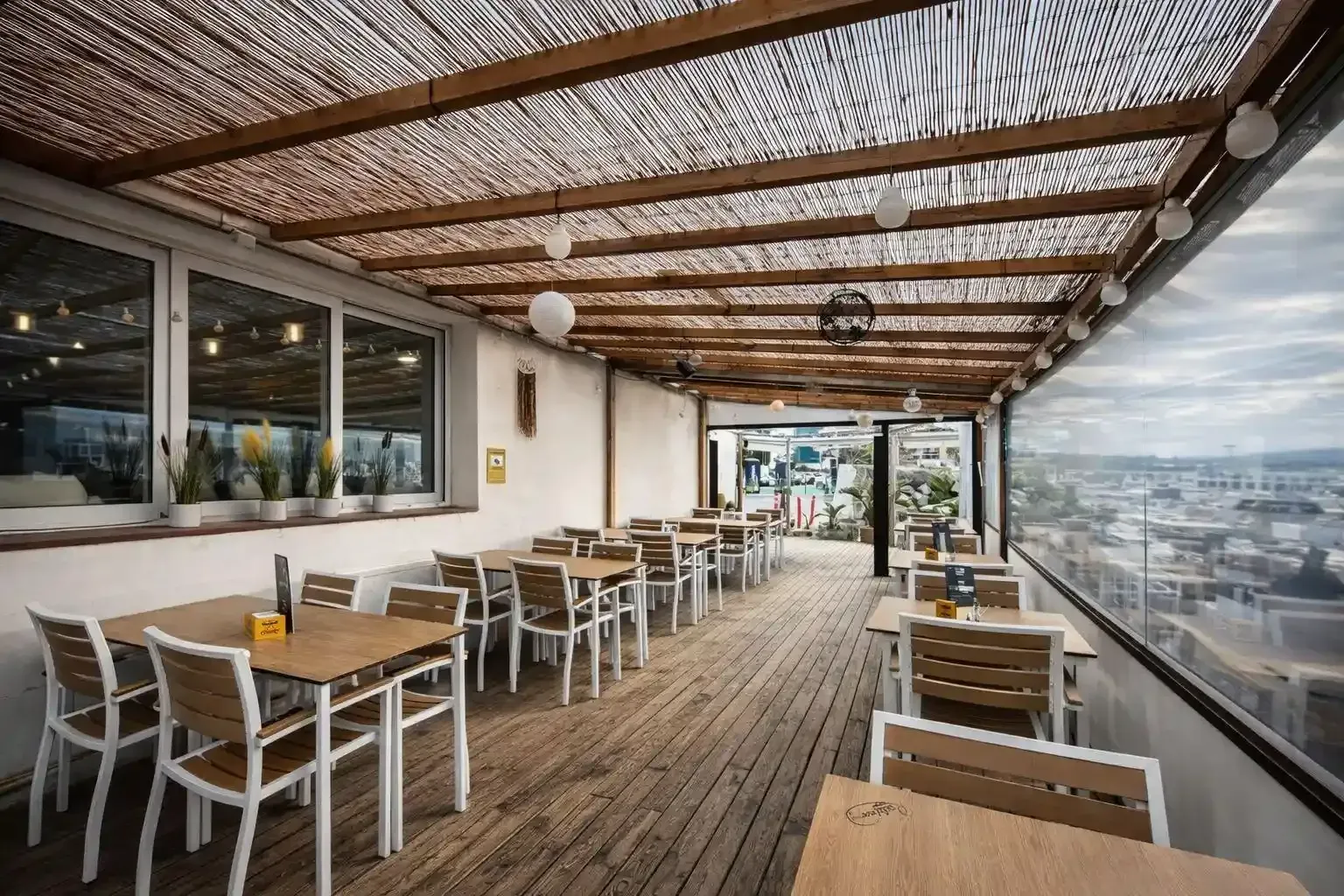 Terraza de restaurante al aire libre con mesas y sillas de madera y vistas a la ciudad bajo un techo de lamas.