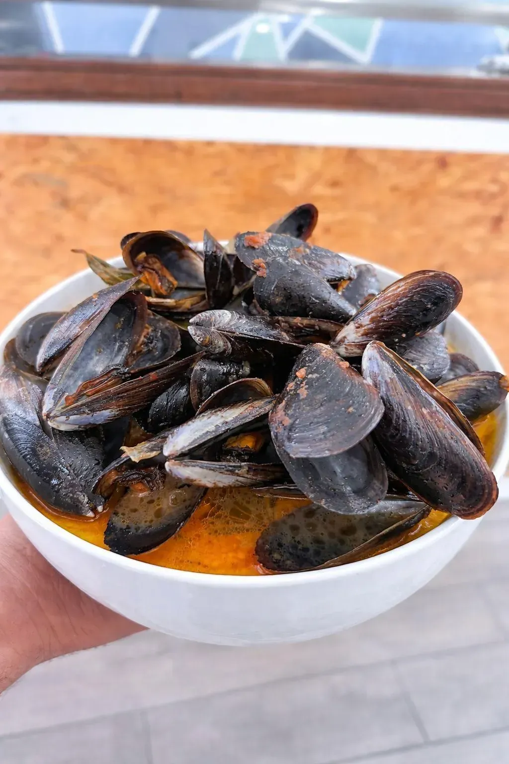 Un bol de mejillones al vapor en caldo de naranja, sostenido en una mano al aire libre.