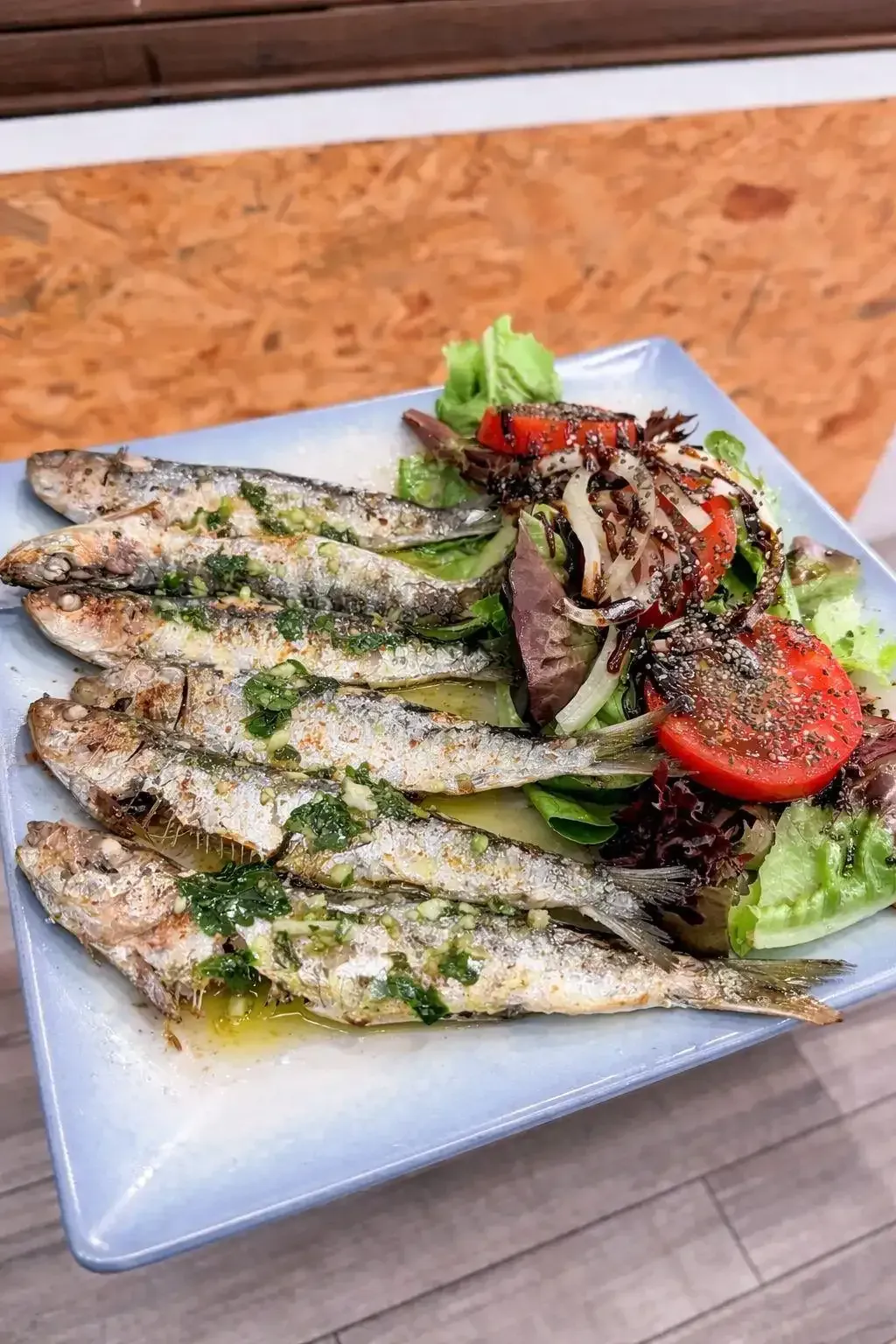 Pescado a la parrilla con costra de hierbas, servido en un plato cuadrado con ensalada mixta y rodajas de tomate.