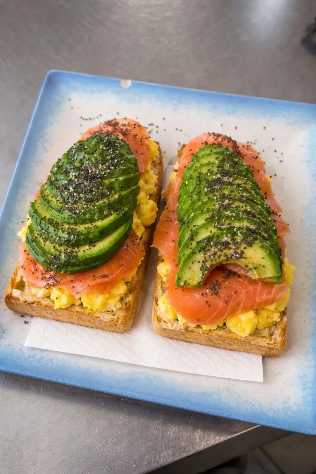 Tostada de aguacate con salmón ahumado y huevos revueltos en un plato con borde azul.