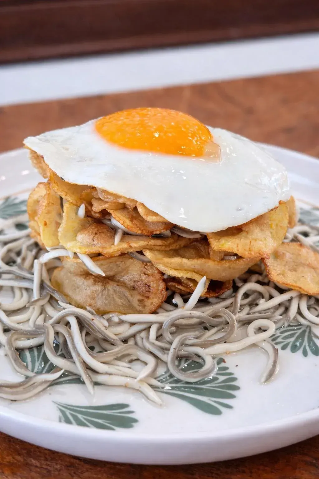 Fideos cubiertos con huevo frito y rodajas de patata crujientes en un plato decorado con flores.
