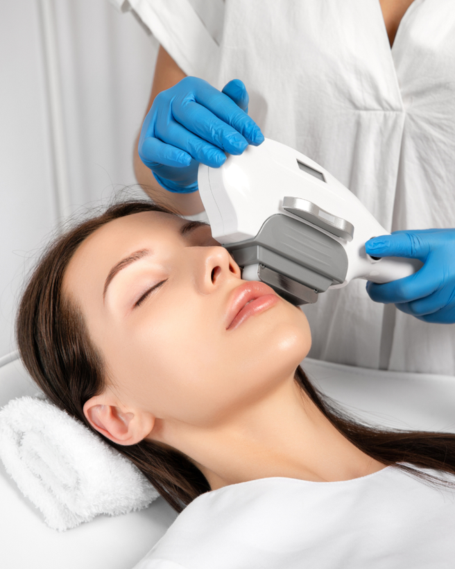 Una profesional con guantes azules utiliza un dispositivo cosmético manual para realizar un tratamiento facial en el rostro de una clienta.