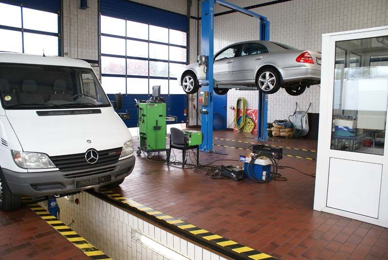 In einer Garage steht auf einer Hebebühne ein weißer Lieferwagen neben einem silbernen Auto.