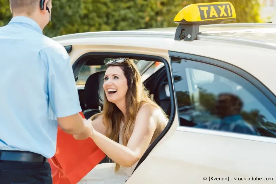 Ein Fahrer in einem blauen Hemd überreicht einem lächelnden Fahrgast in einem Taxi eine rote Einkaufstasche.