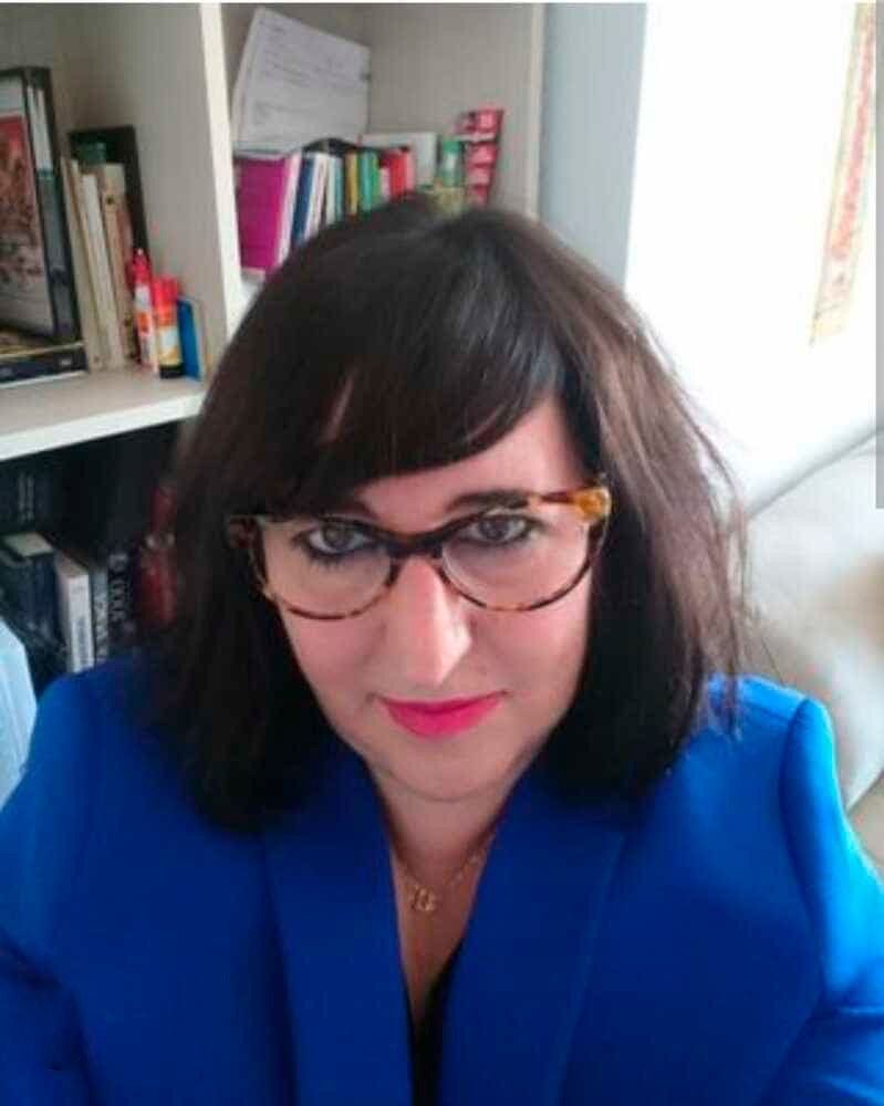 Una mujer que lleva gafas y una chaqueta azul está sentada delante de una estantería.