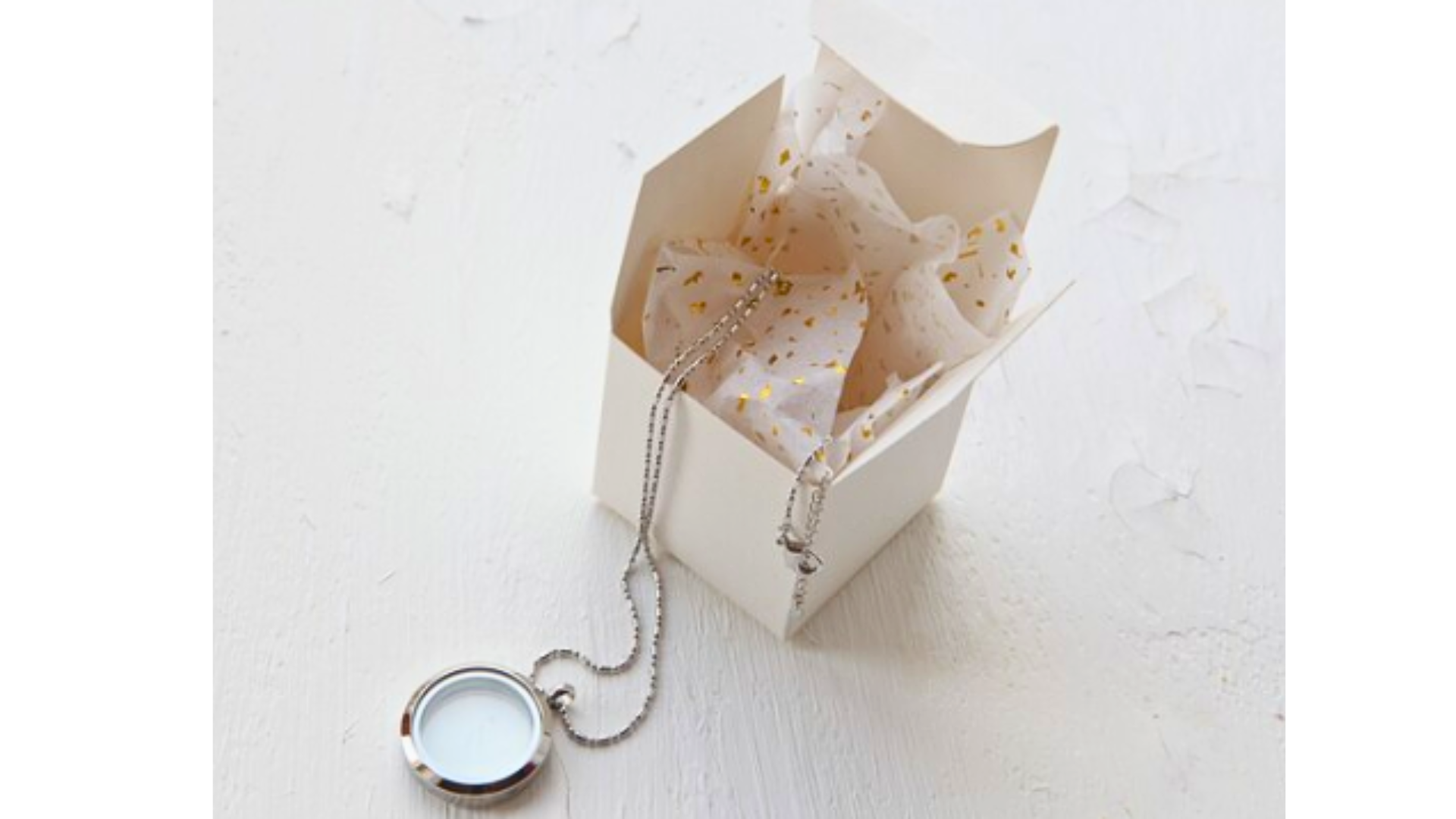 Un collier médaillon en argent dépasse d'une boîte cadeau blanche ouverte, remplie de papier de soie