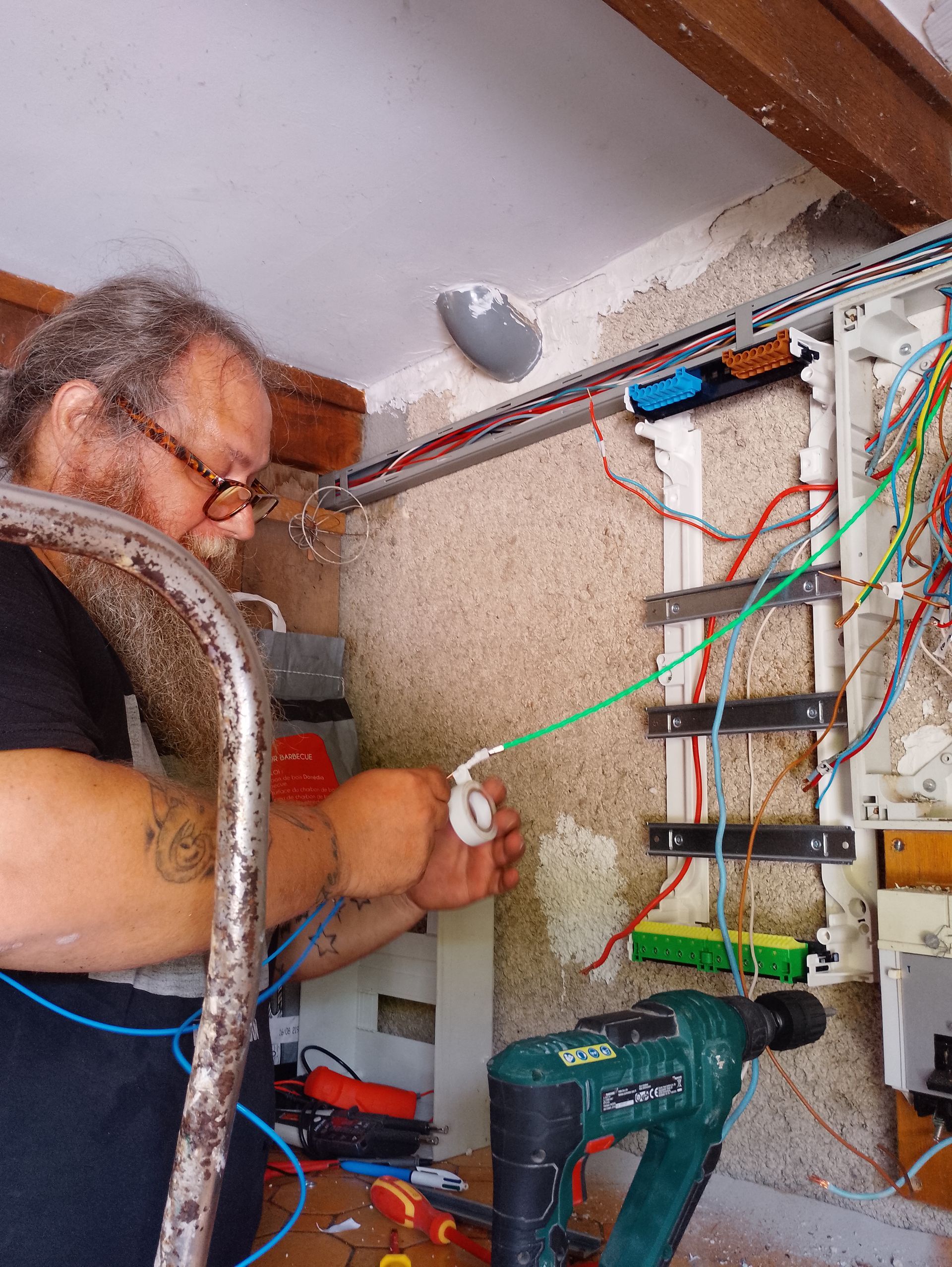 Un électricien installe un tableau électrique avec des câbles à côté d'une perceuse dans un local technique.