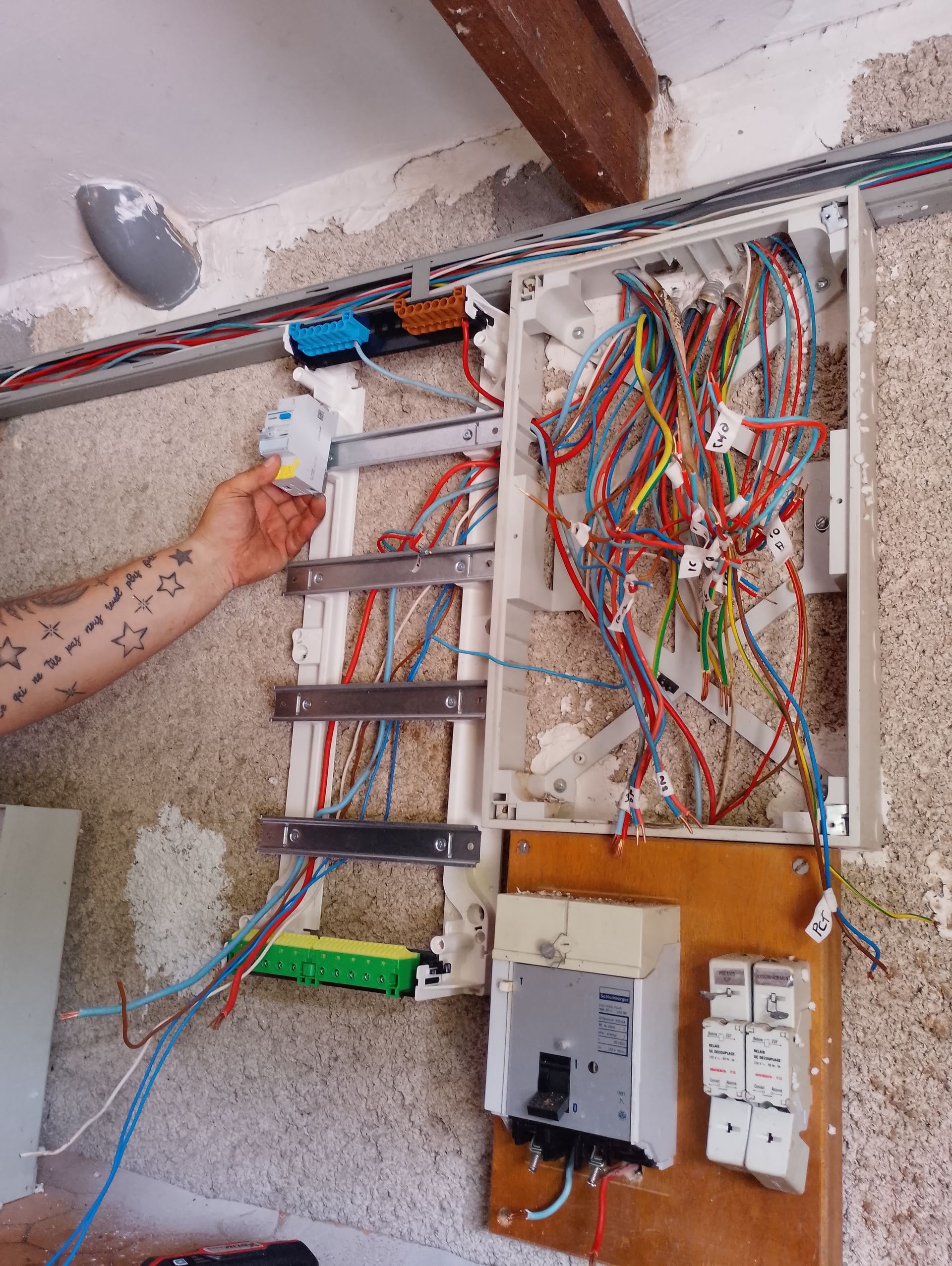 Tableau électrique ouvert avec des fils apparents et une main pointant vers un bloc de fusibles sur un mur rugueux.