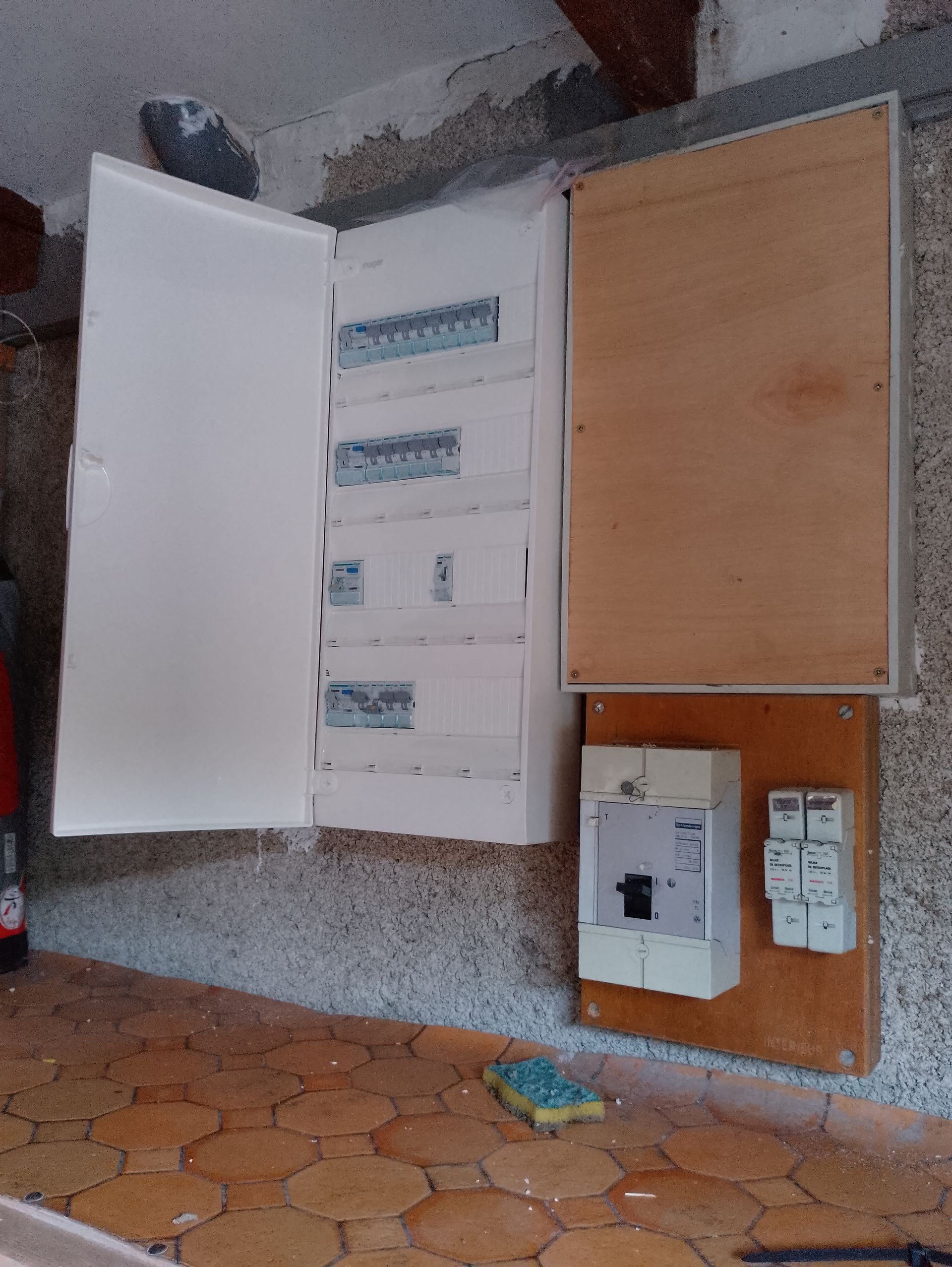 Tableau électrique ouvert avec disjoncteurs apparents dans un coin de mur moucheté au-dessus d'un sol carrelé.