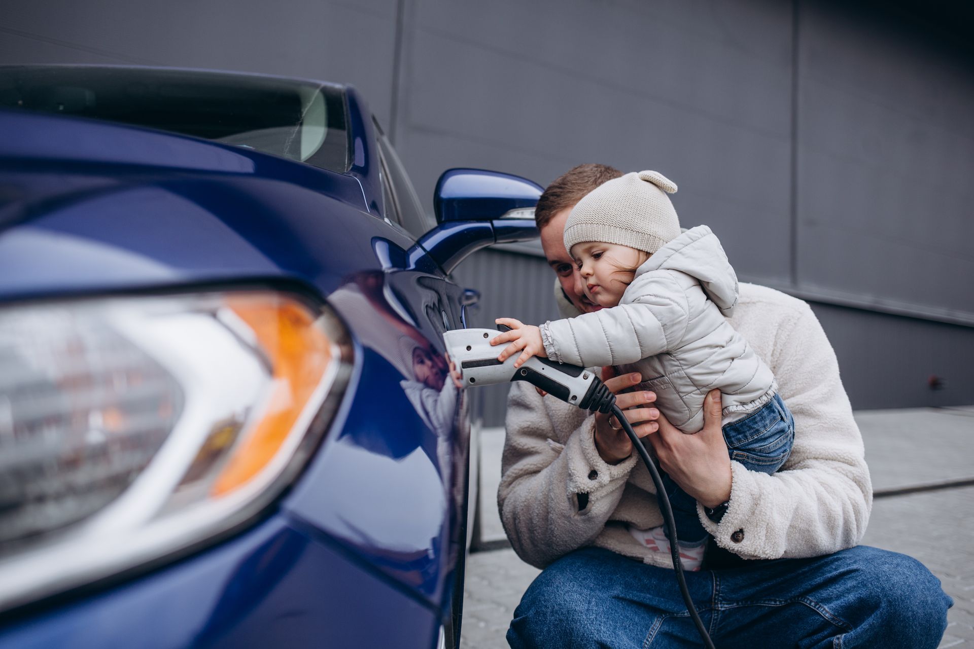 Une femme accroupie près d'une voiture bleue, tenant un enfant près de la portière ouverte.