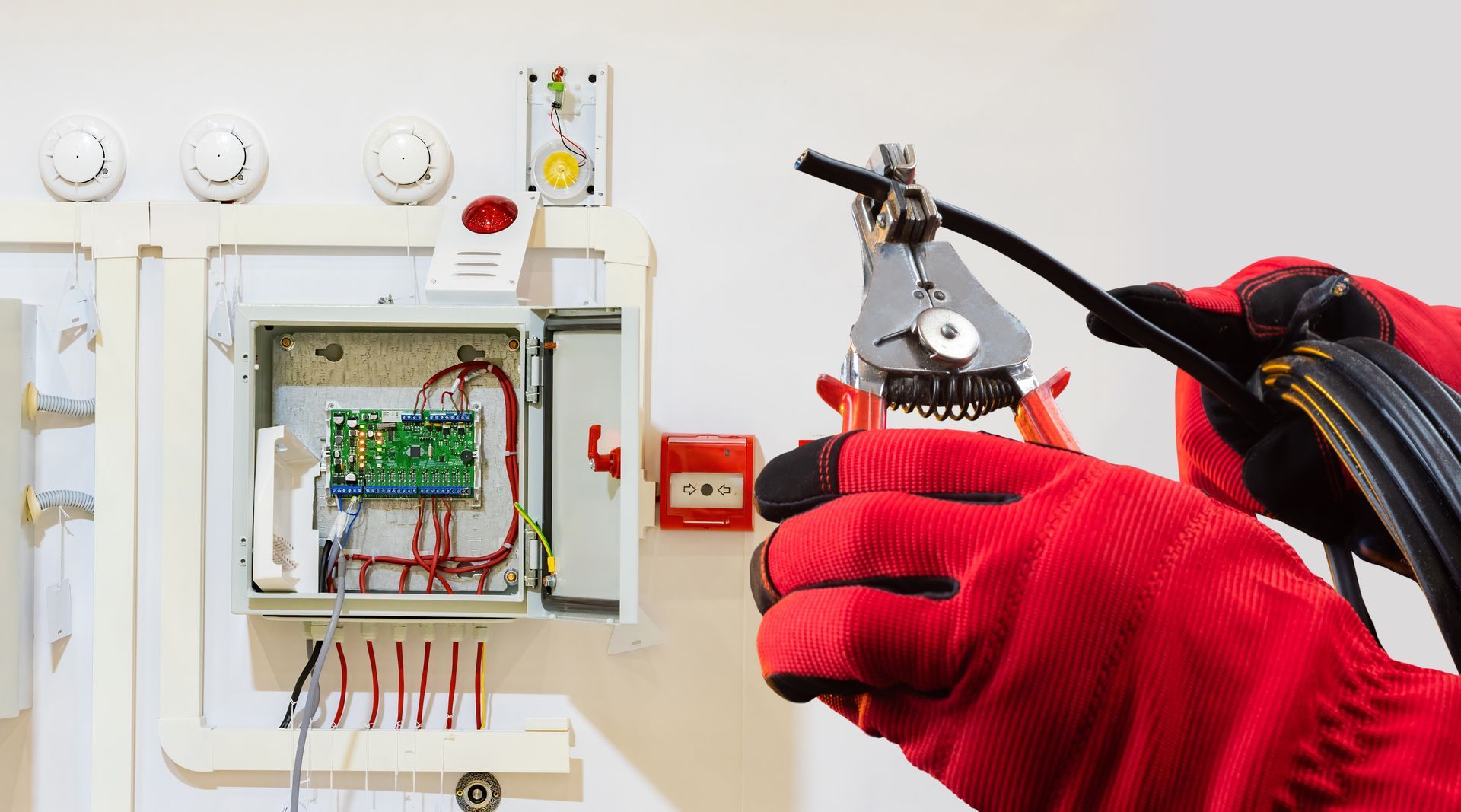 Des mains gantées de rouge utilisent un multimètre sur les fils électriques apparents d'un panneau mural.