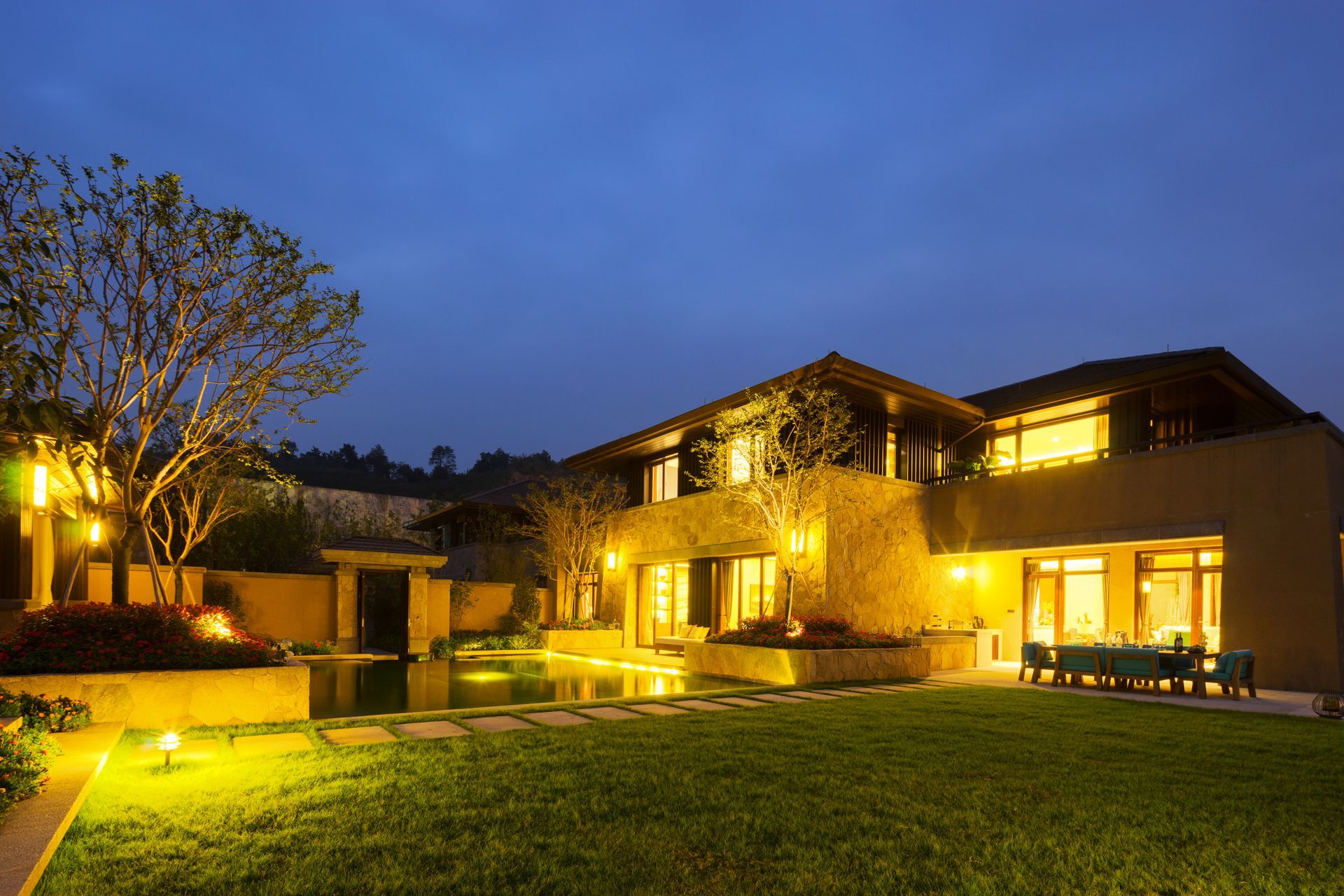 Maison moderne illuminée et pelouse paysagée au crépuscule, avec des lumières chaudes et une terrasse éclairée.