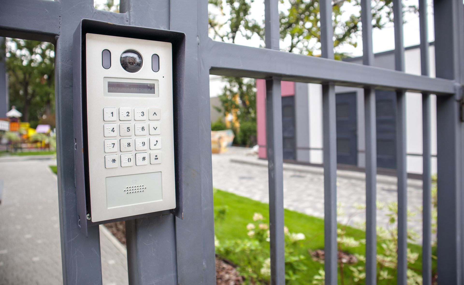 Interphone à clavier métallique fixé sur un portail gris à côté d'un trottoir et de l'entrée de l'immeuble.