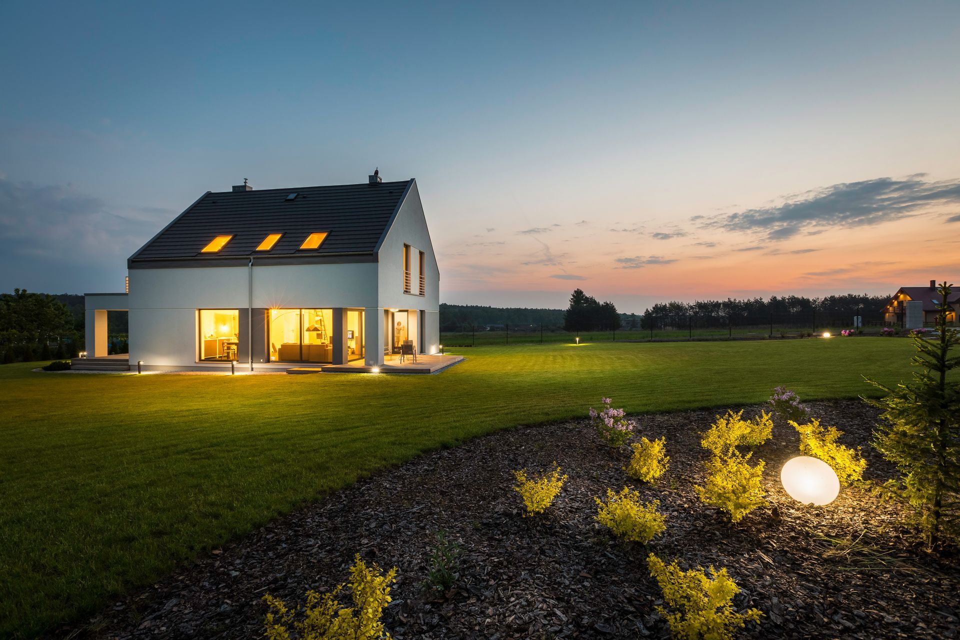 Maison blanche illuminée au crépuscule, à côté d'une pelouse verte, avec une lampe de jardin scintillante au premier plan.