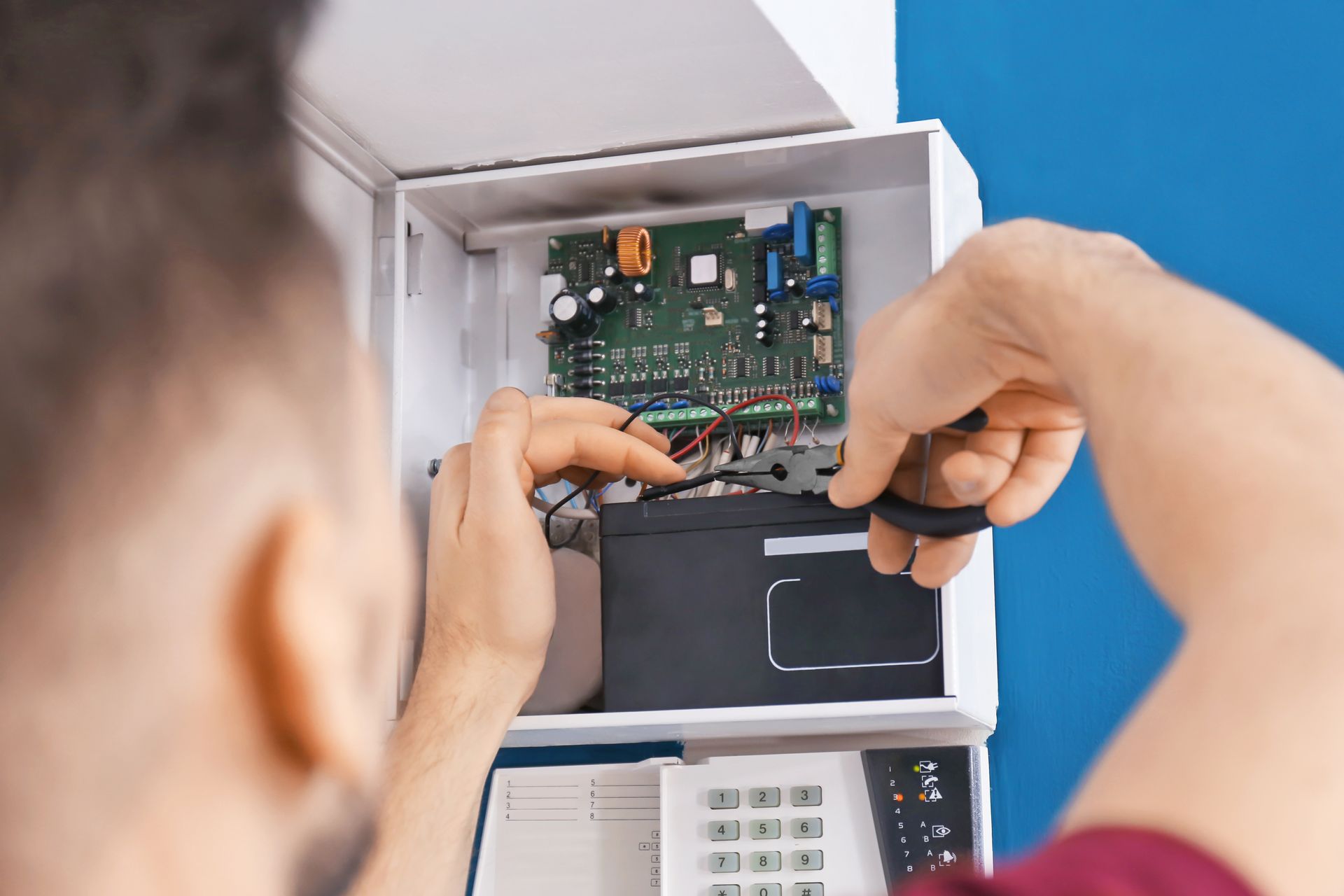 Technicien réparant un panneau de commande d'alarme ouvert, avec circuit imprimé et clavier exposés.
