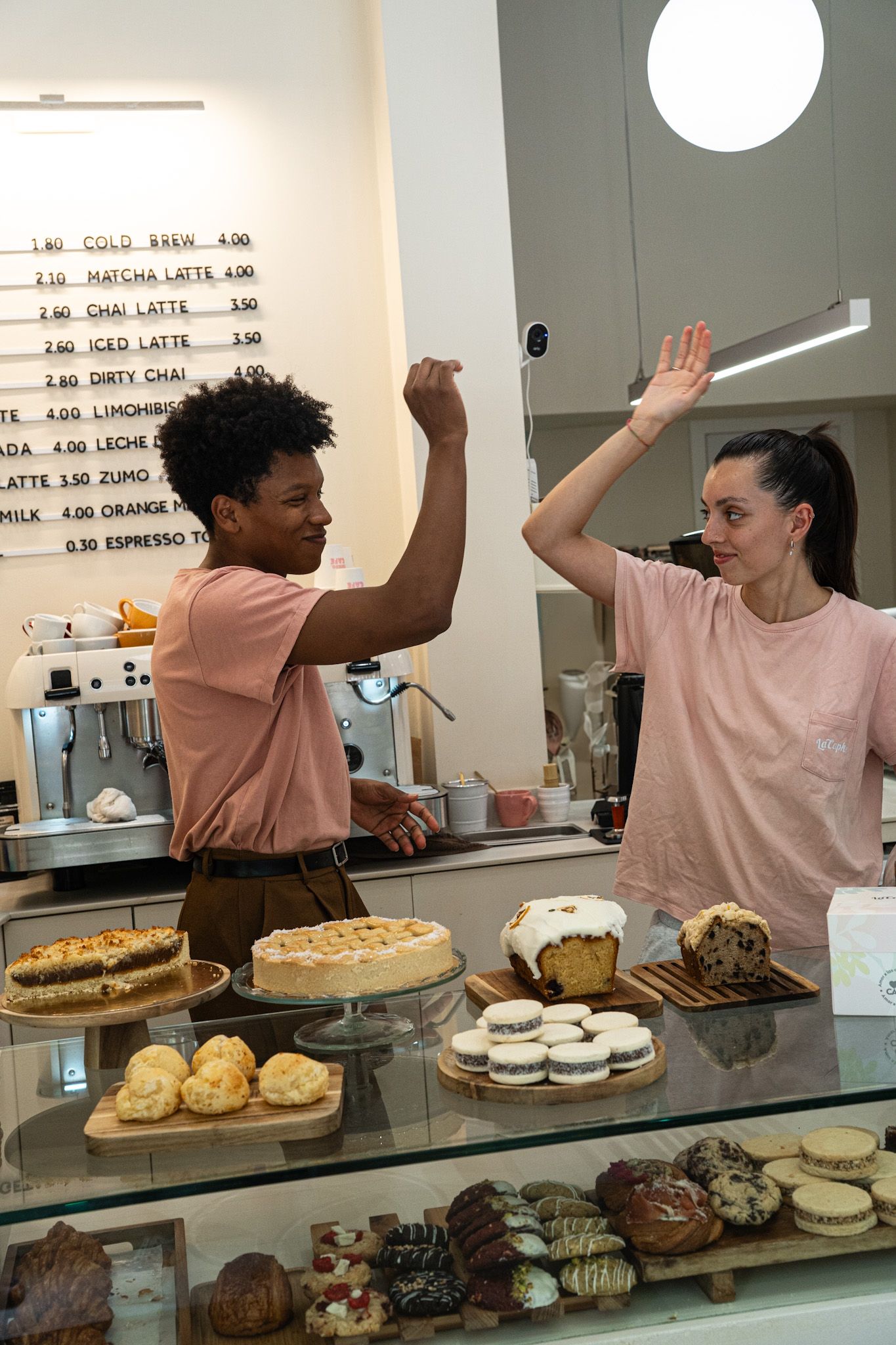 Dos personas con camisetas rosas chocan las manos detrás de un mostrador de una pastelería lleno de pasteles y bollería.