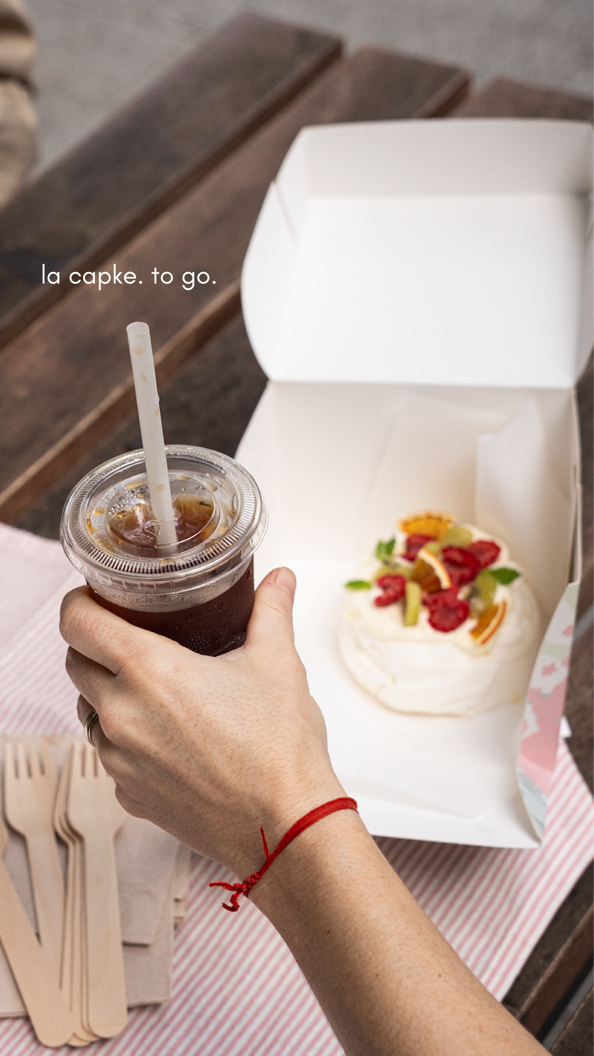 Una mano sostiene un café helado junto a un pastel en una caja blanca abierta sobre una mesa de madera.