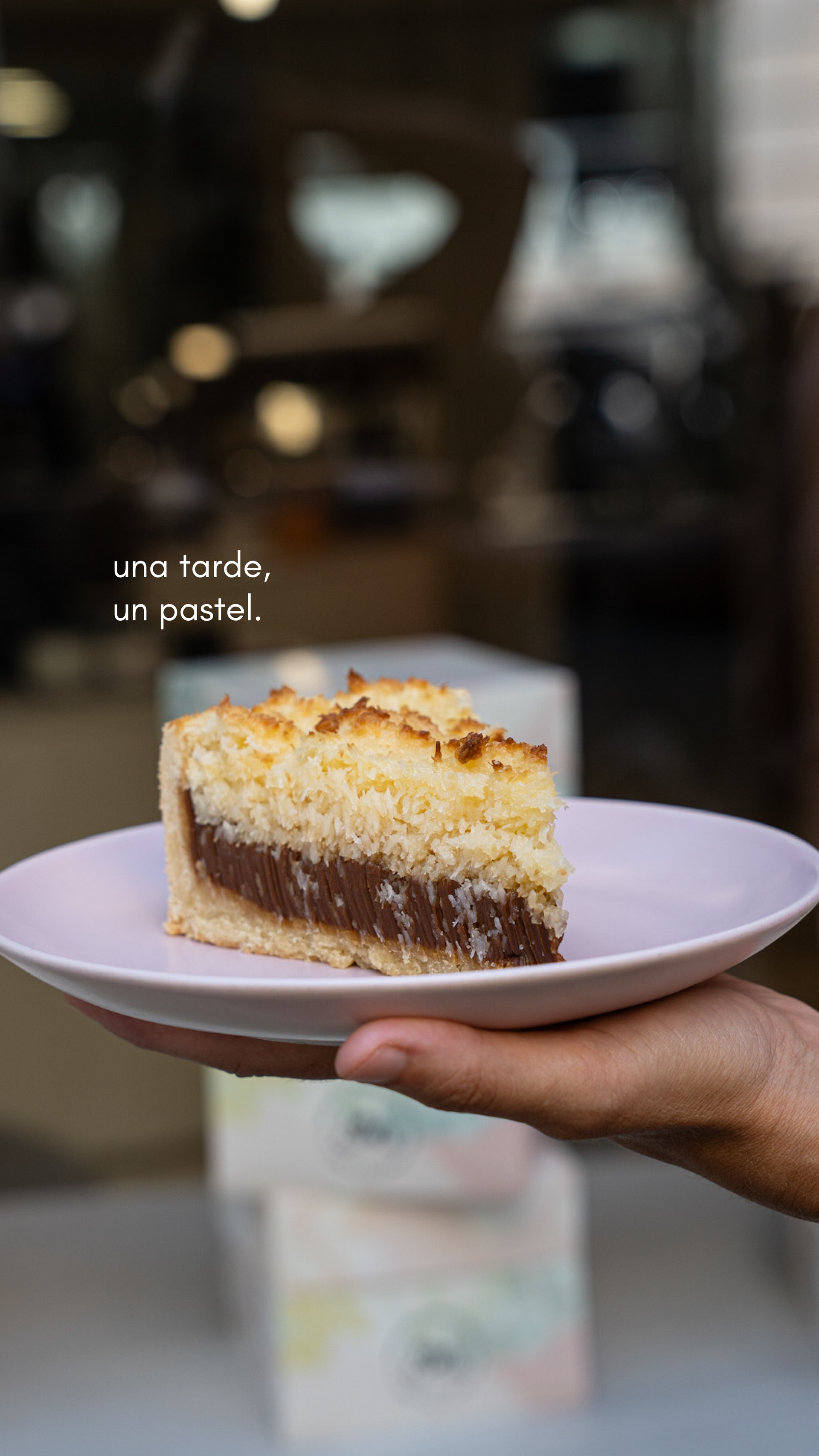 Una porción de pastel de varias capas sobre un plato blanco, sostenida por una mano contra el fondo borroso de una cafetería.