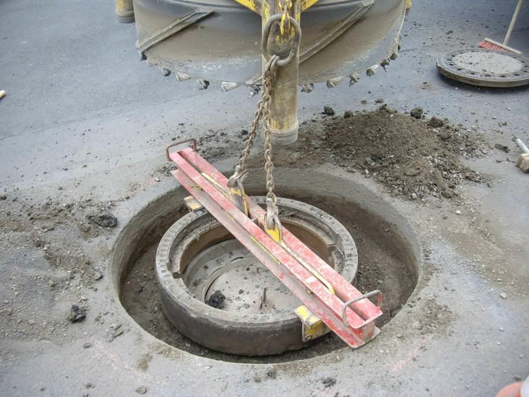 Ein Schachtdeckel wird von einem Kran angehoben, an dem eine rote Wasserwaage befestigt ist