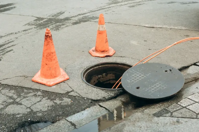 Conos naranjas junto a un pozo abierto sobre asfalto agrietado, con cables visibles.