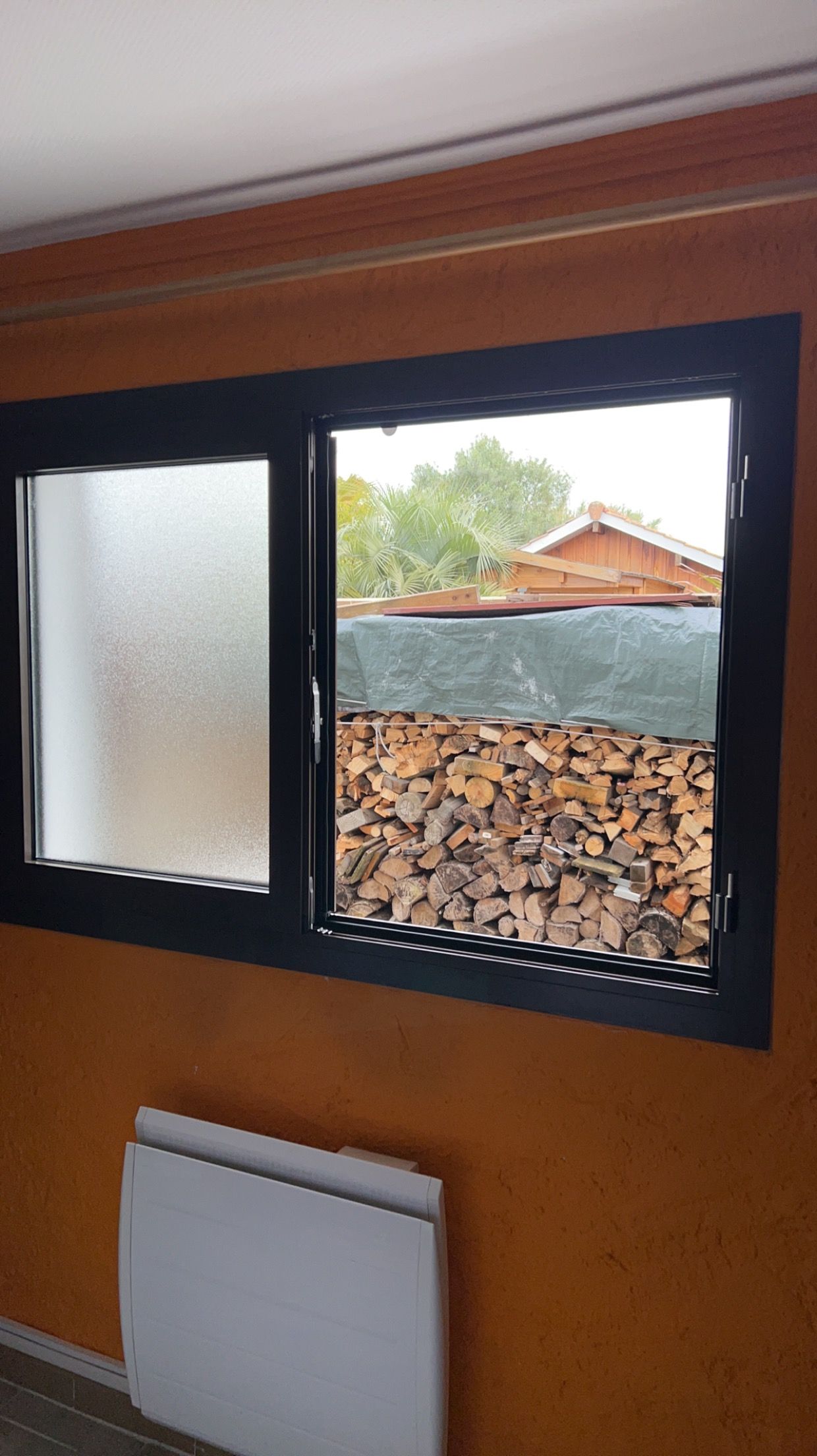 Vue intérieure d'une fenêtre à verre dépoli, bois de chauffage empilé à l'extérieur et radiateur mural blanc en dessous.