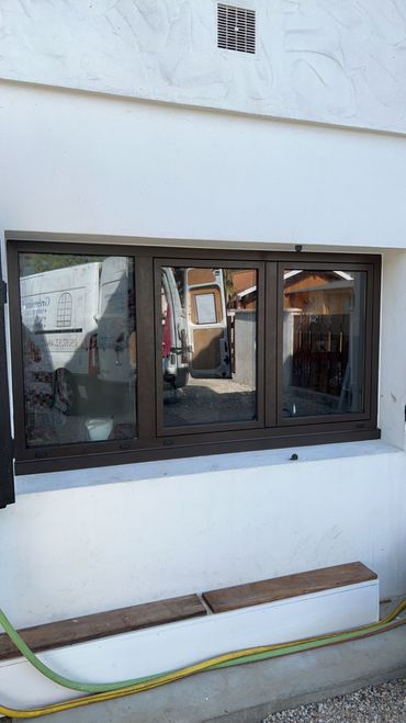 Petite fenêtre à cadre noir dans un mur extérieur blanc, avec un tuyau d'arrosage et un banc en dessous.