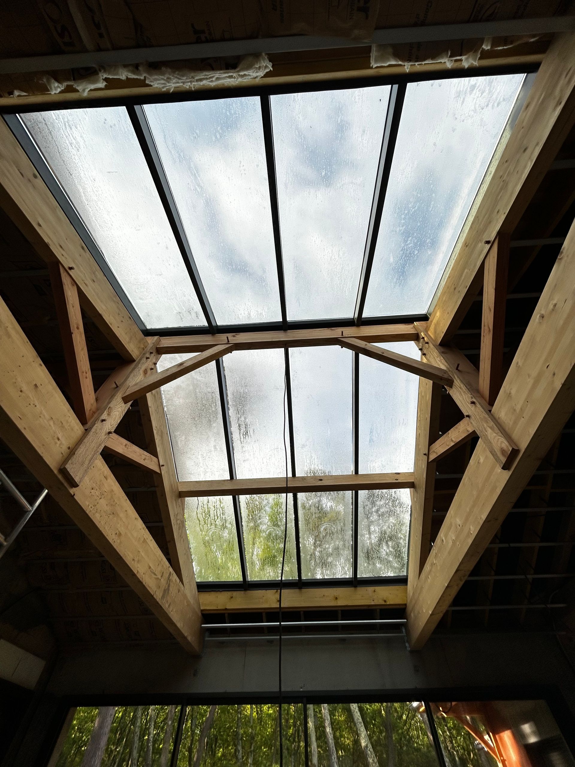 Vue intérieure d'une charpente en bois avec une grande lucarne et un ciel nuageux au-dessus