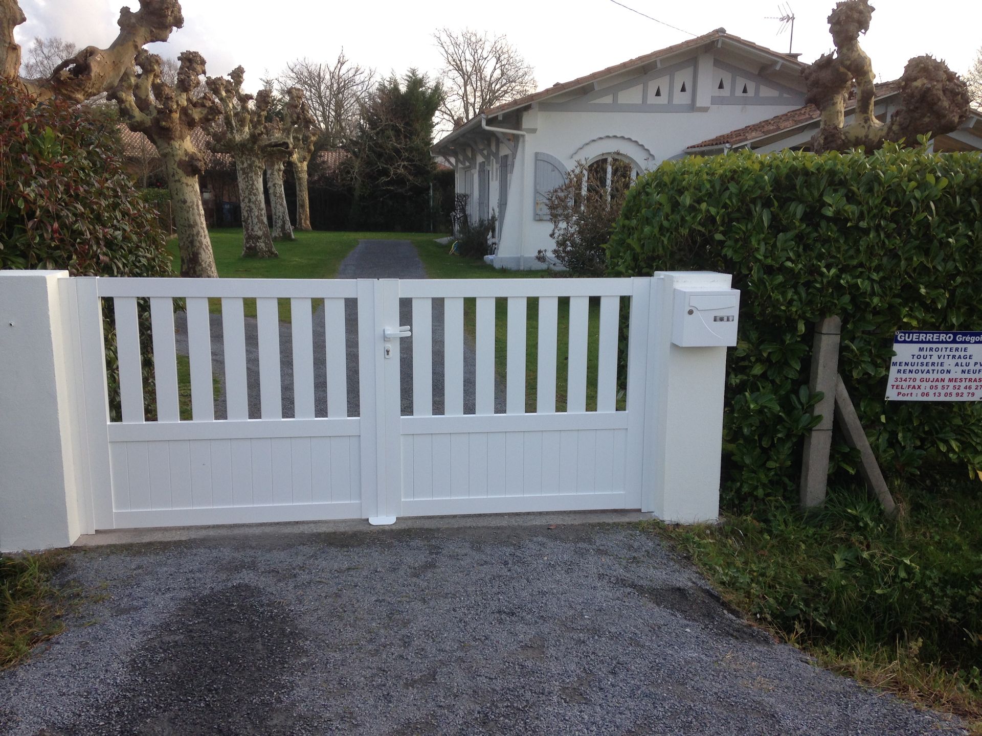 Portail blanc devant une maison avec un chemin de gravier et des haies au crépuscule