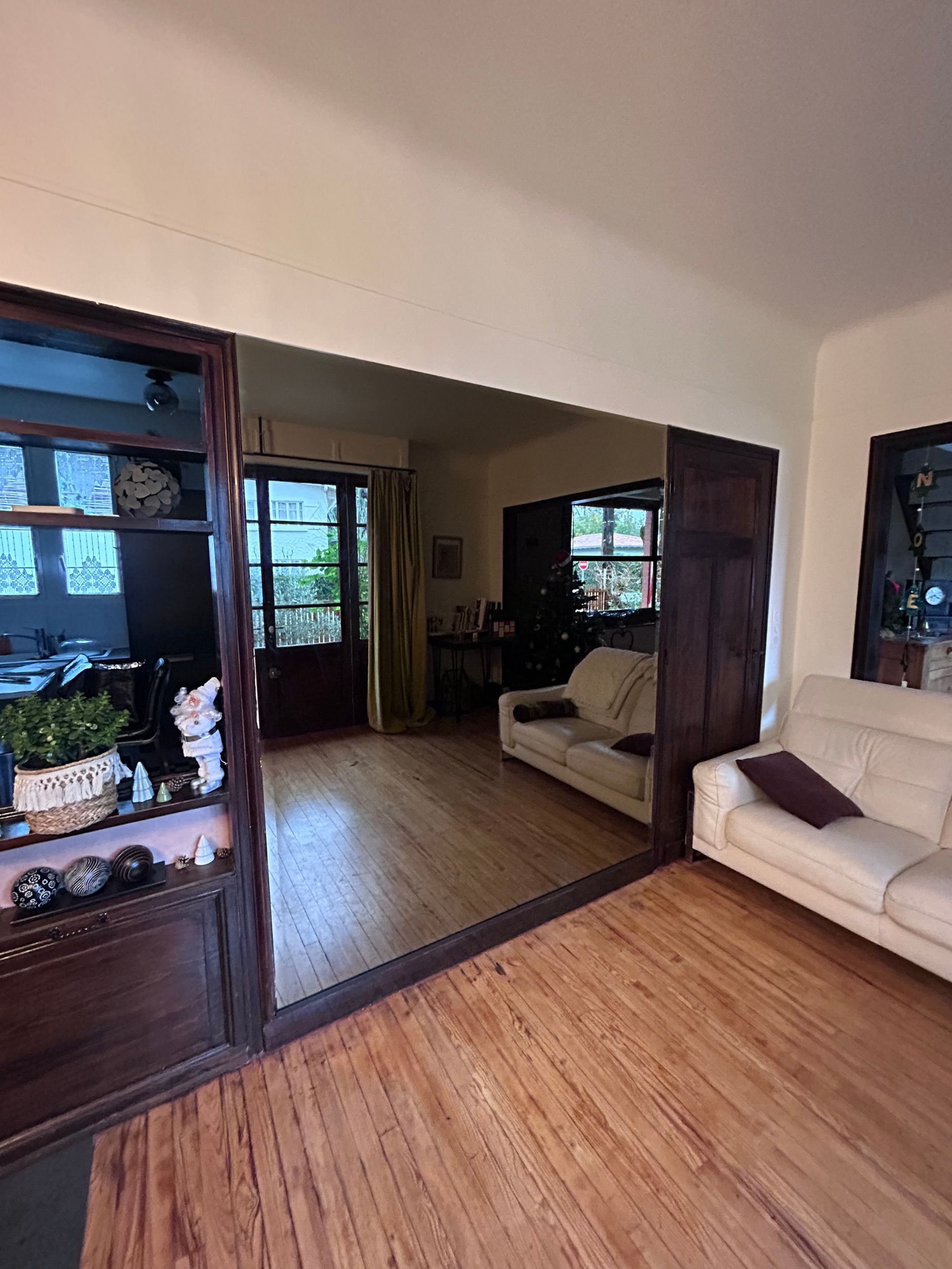 Salon avec parquet, grand mur de miroirs, canapé beige et fenêtres donnant sur la verdure.