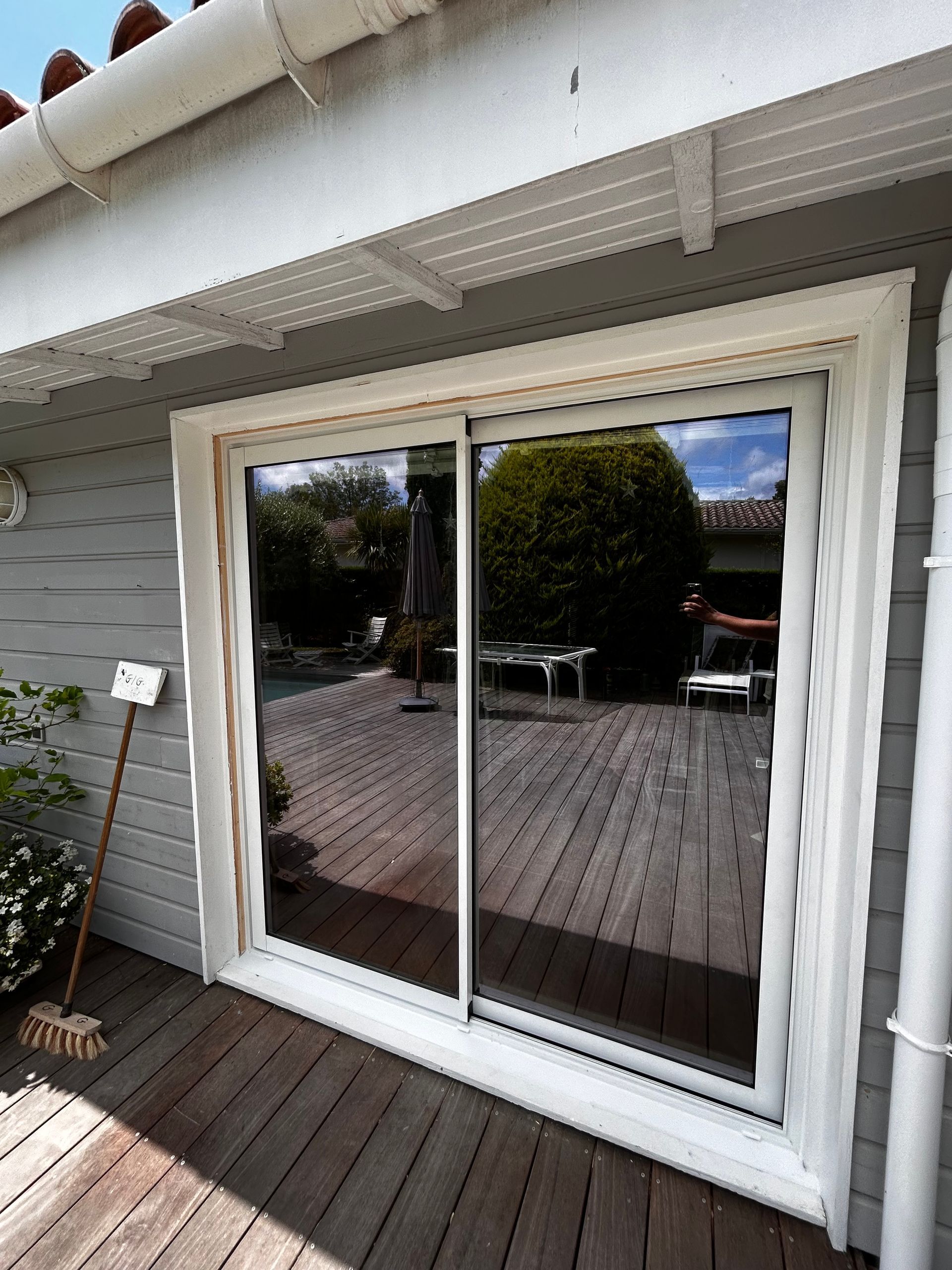 Porte coulissante en verre blanc avec encadrement beige sur une maison grise, reflétant une terrasse en bois et un jardin.