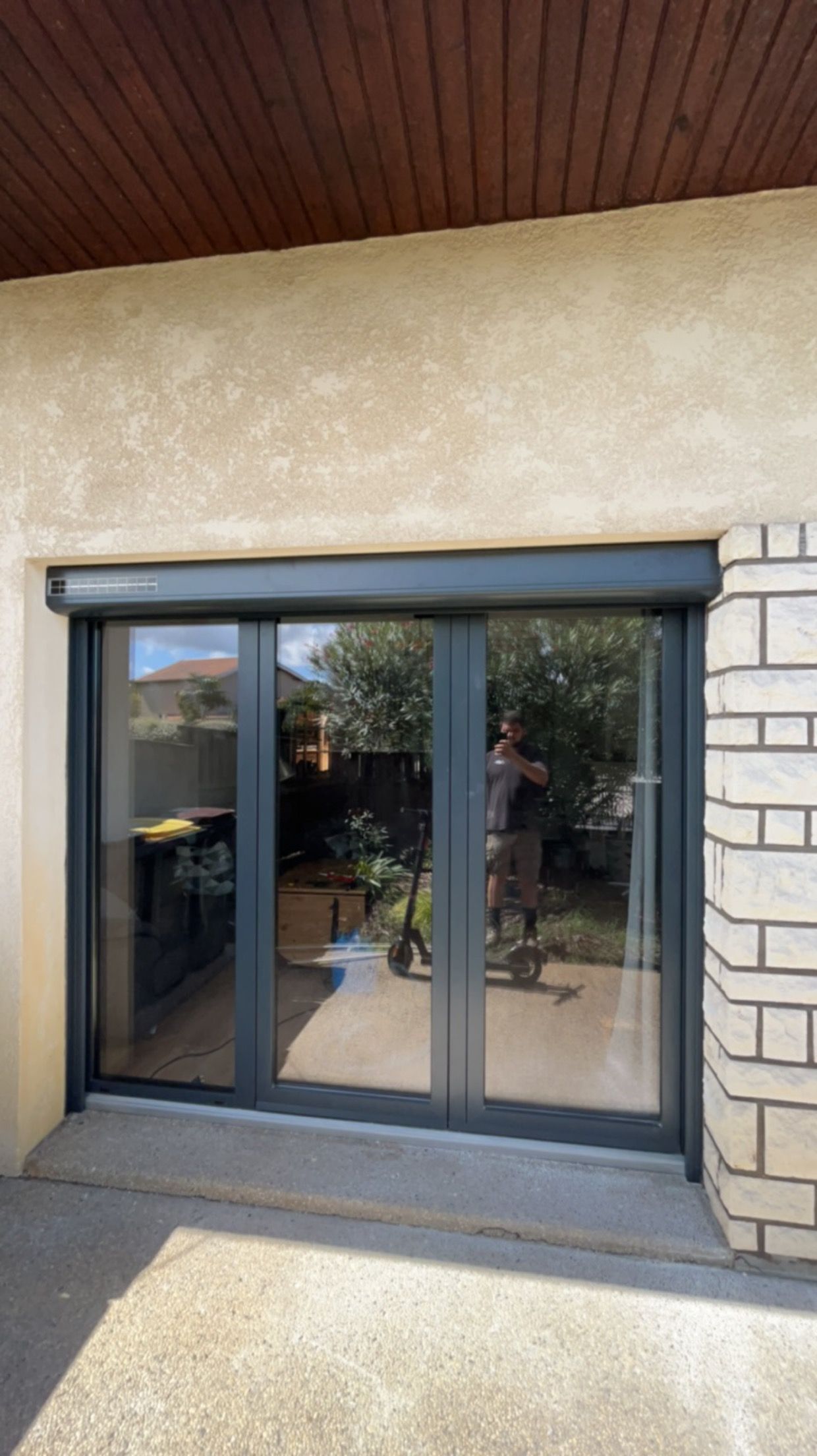 Porte-fenêtre coulissante en verre intégrée à un mur extérieur beige, avec un auvent couvert et un panneau latéral en briques.