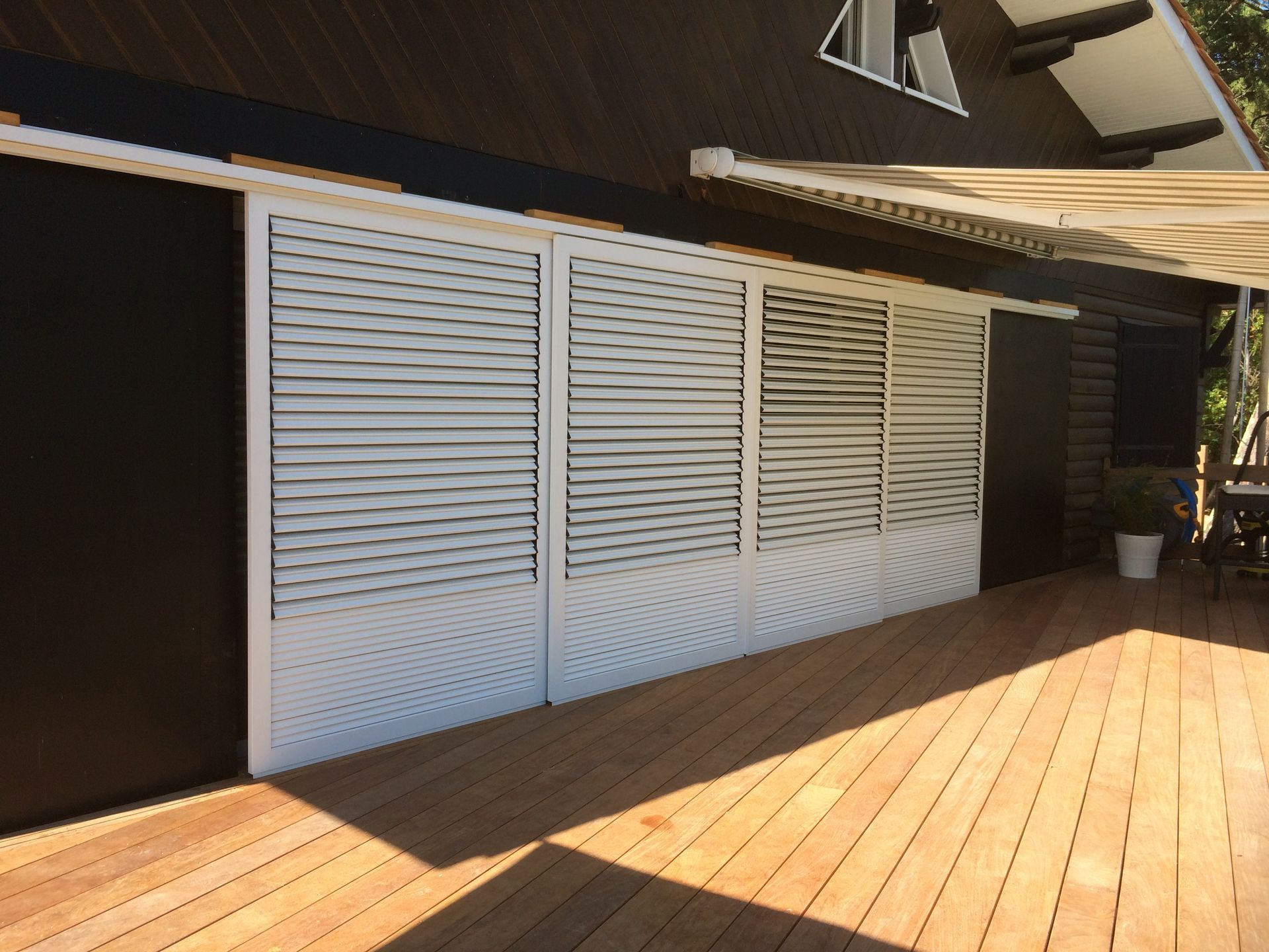 Terrasse en bois longeant un mur blanc à persiennes, jouant avec les ombres du soleil et abritant un avant-toit sombre.