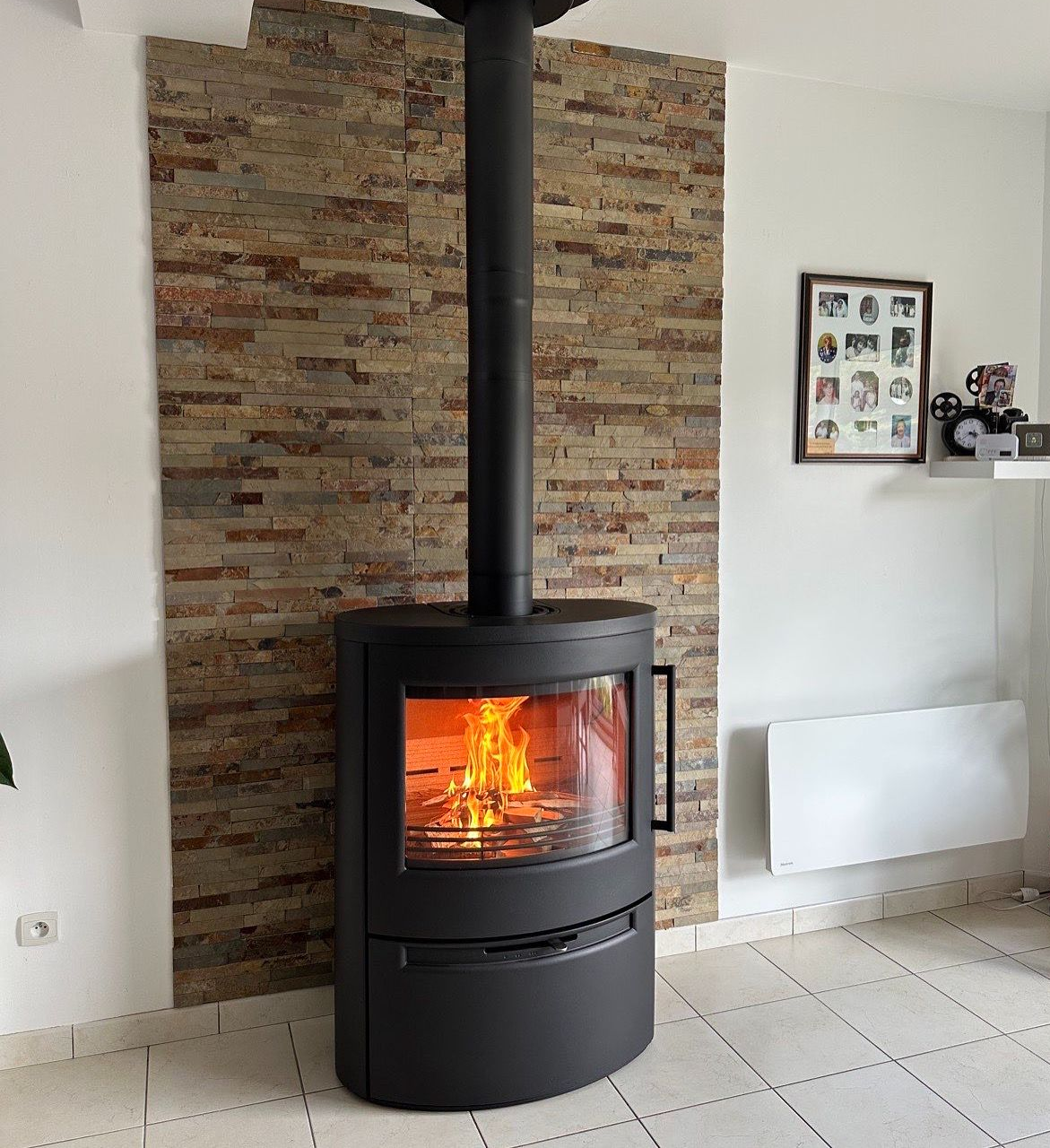 Poêle à bois noir moderne avec porte vitrée et flammes visibles sur un mur lambrissé clair.