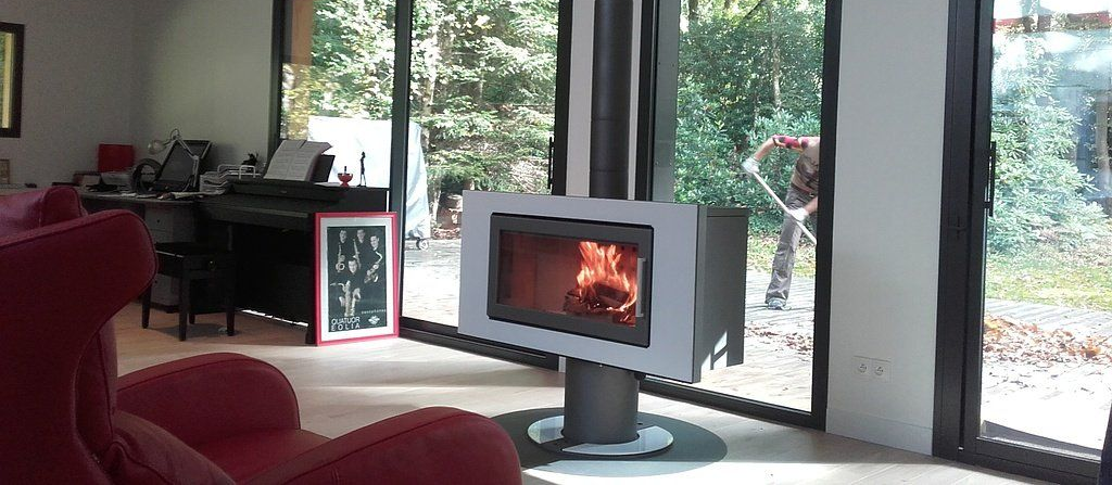 Salon chaleureux avec un poêle à bois allumé, des bûches empilées, un fauteuil en osier et un tapis rond.